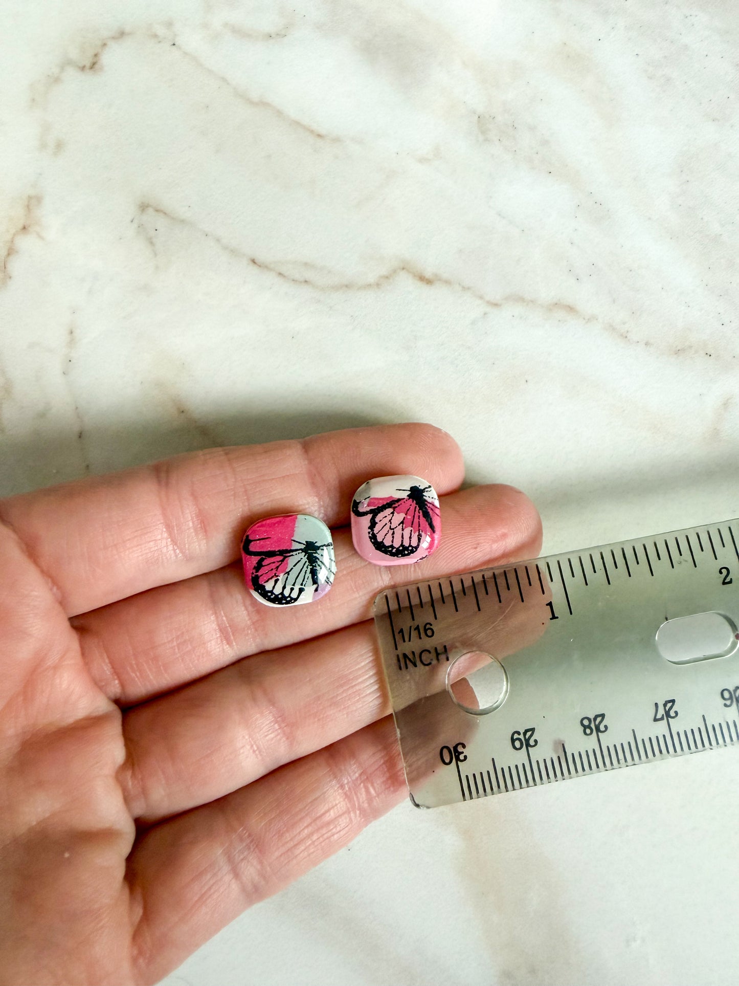 Butterfly-patterned earrings held between fingers with a ruler for scale on a marble background. About a half inch wide 