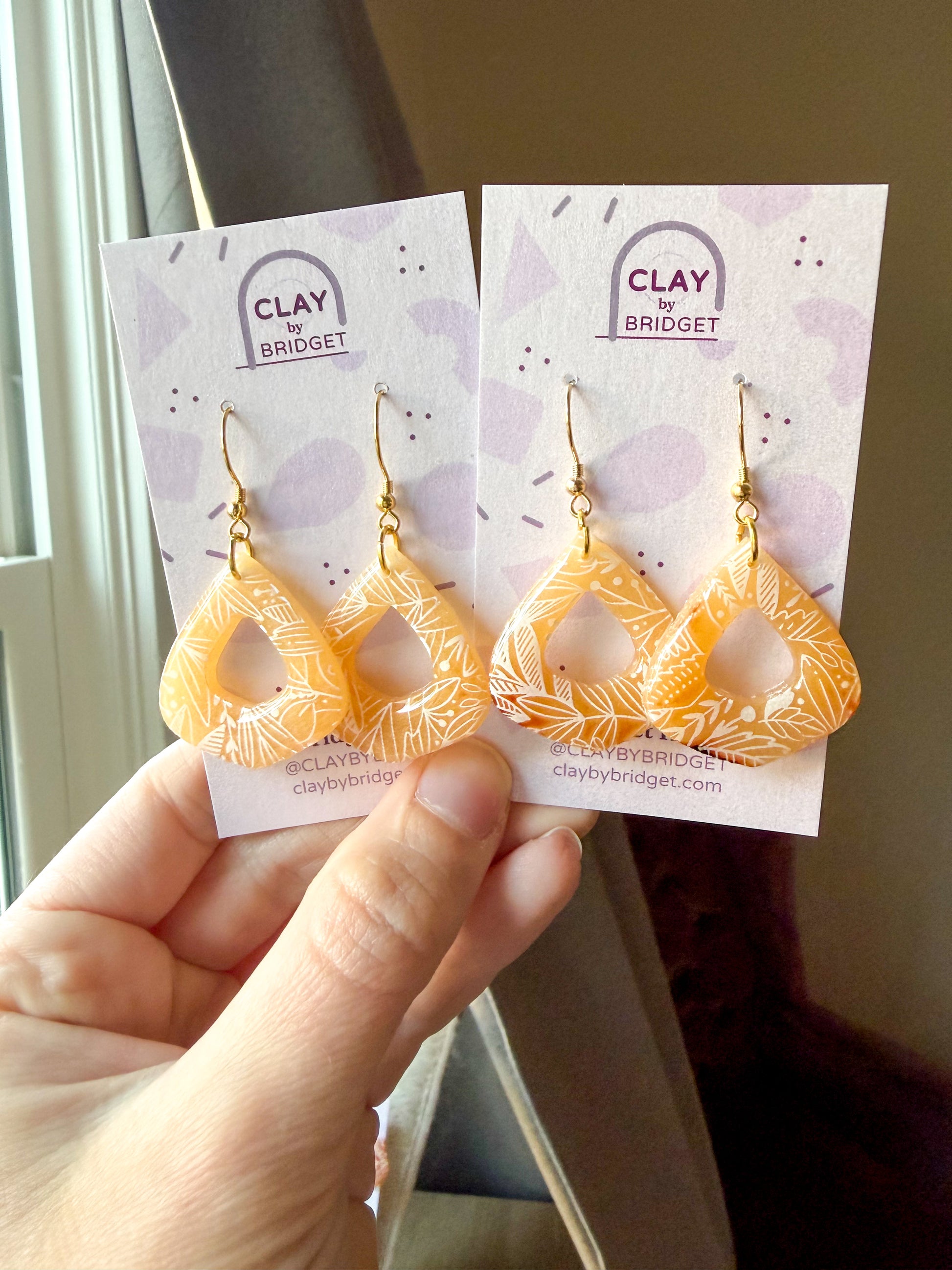 A hand holds two pairs of teardrop-shaped earrings on display cards. The earrings are made of translucent clay with white leaf patterns and gold hooks.