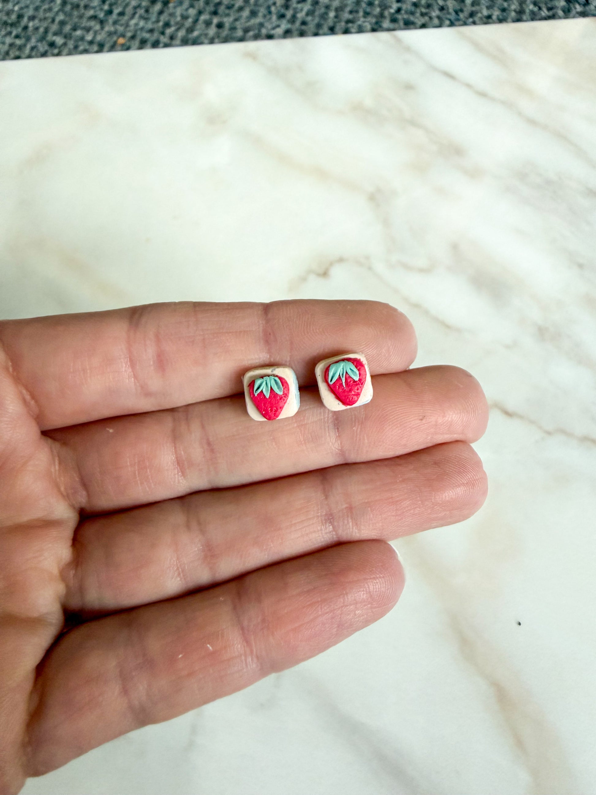 Small strawberry-shaped earrings held between fingers on a marble surface