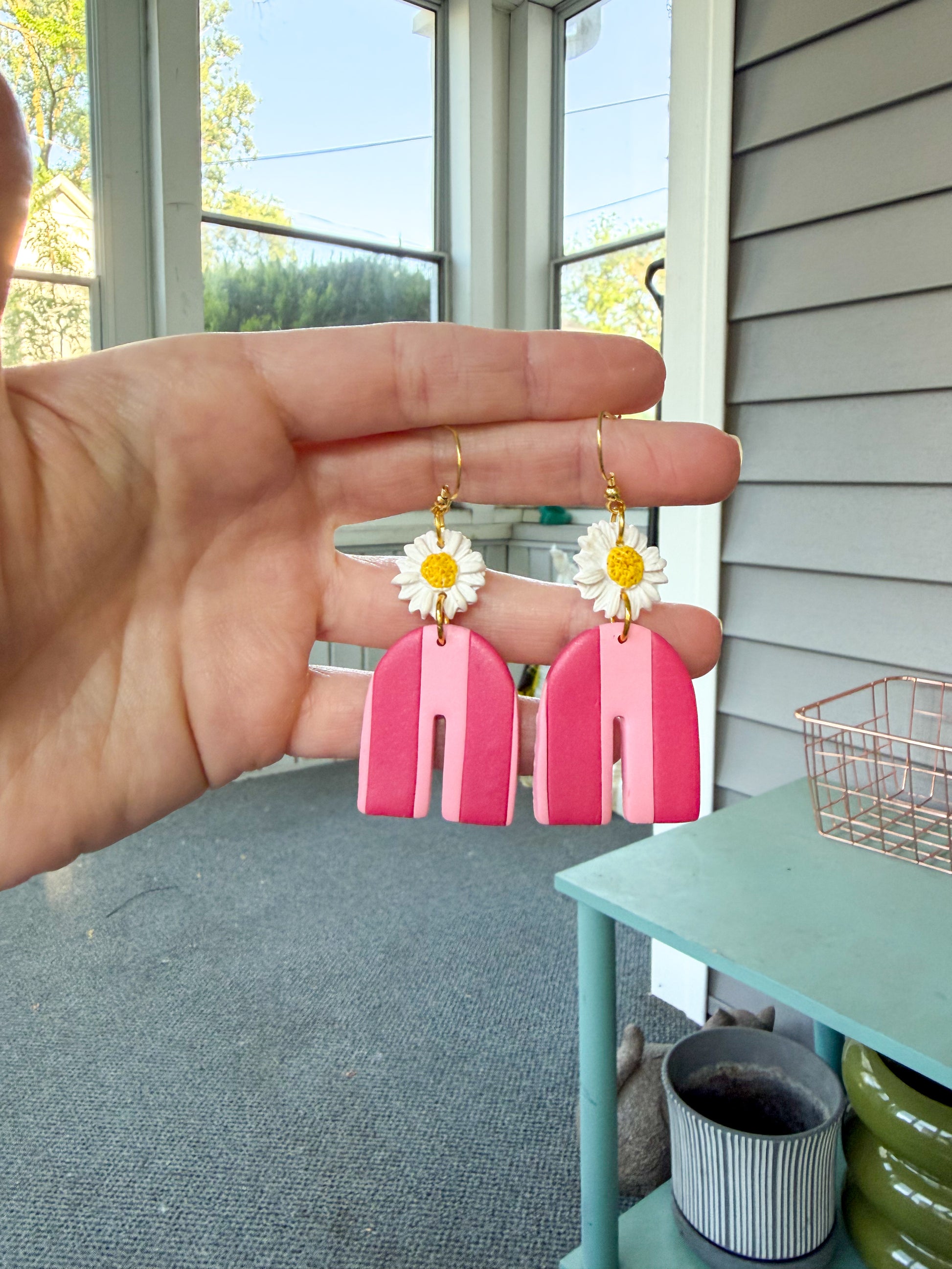 Hand holding pink earrings with floral designs in front of a window.