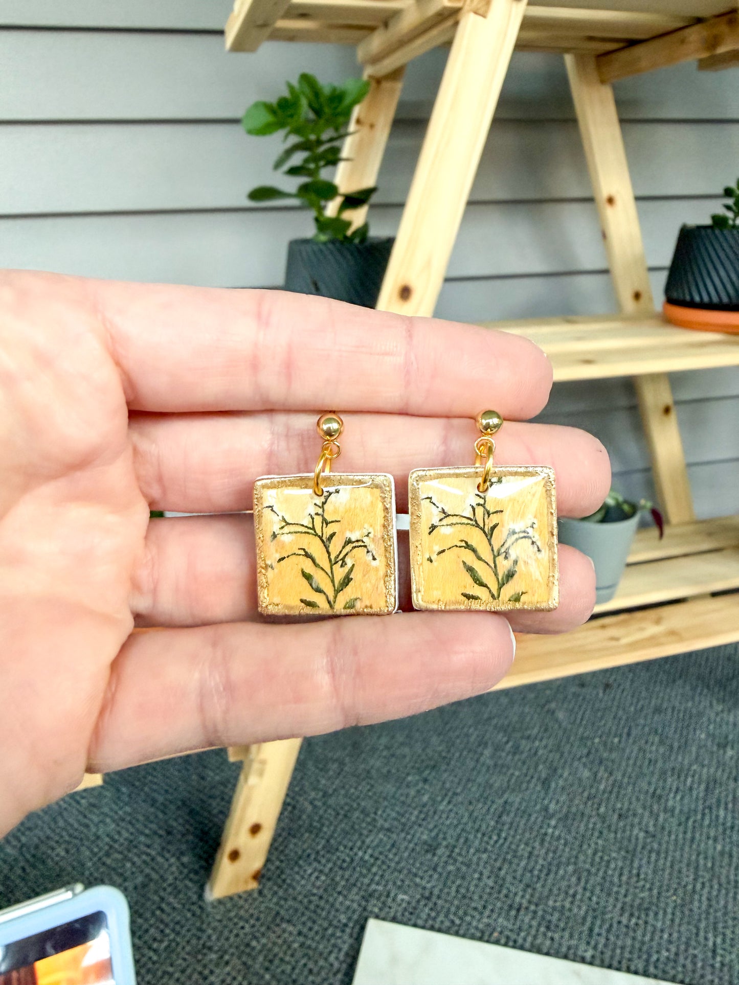 Gold square earrings held in a hand with a wooden chair and plants in the background.
