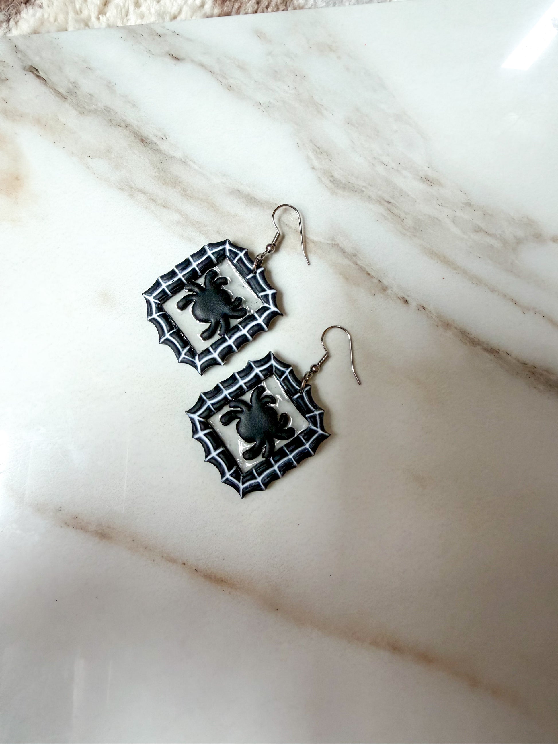 A pair of black and white spider-themed dangle earrings with silver hooks, displayed on a white marble surface.