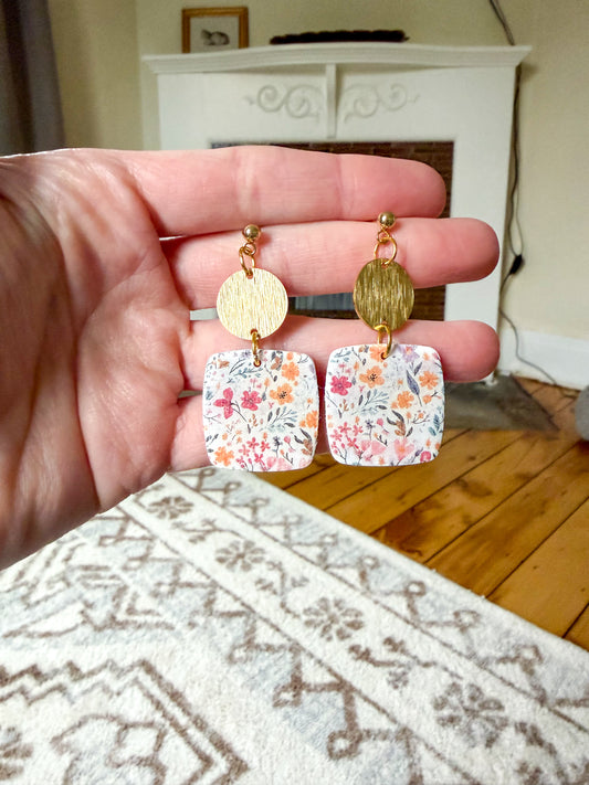 Hand holding floral earrings with gold accents on a wooden floor.