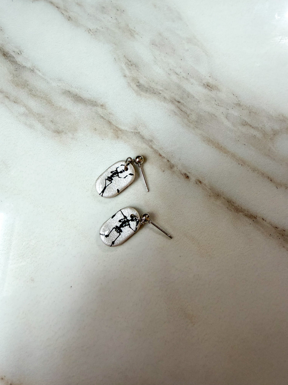 A pair of silver dangle earrings with white oval charms featuring black skeleton designs, resting on a white marble surface.