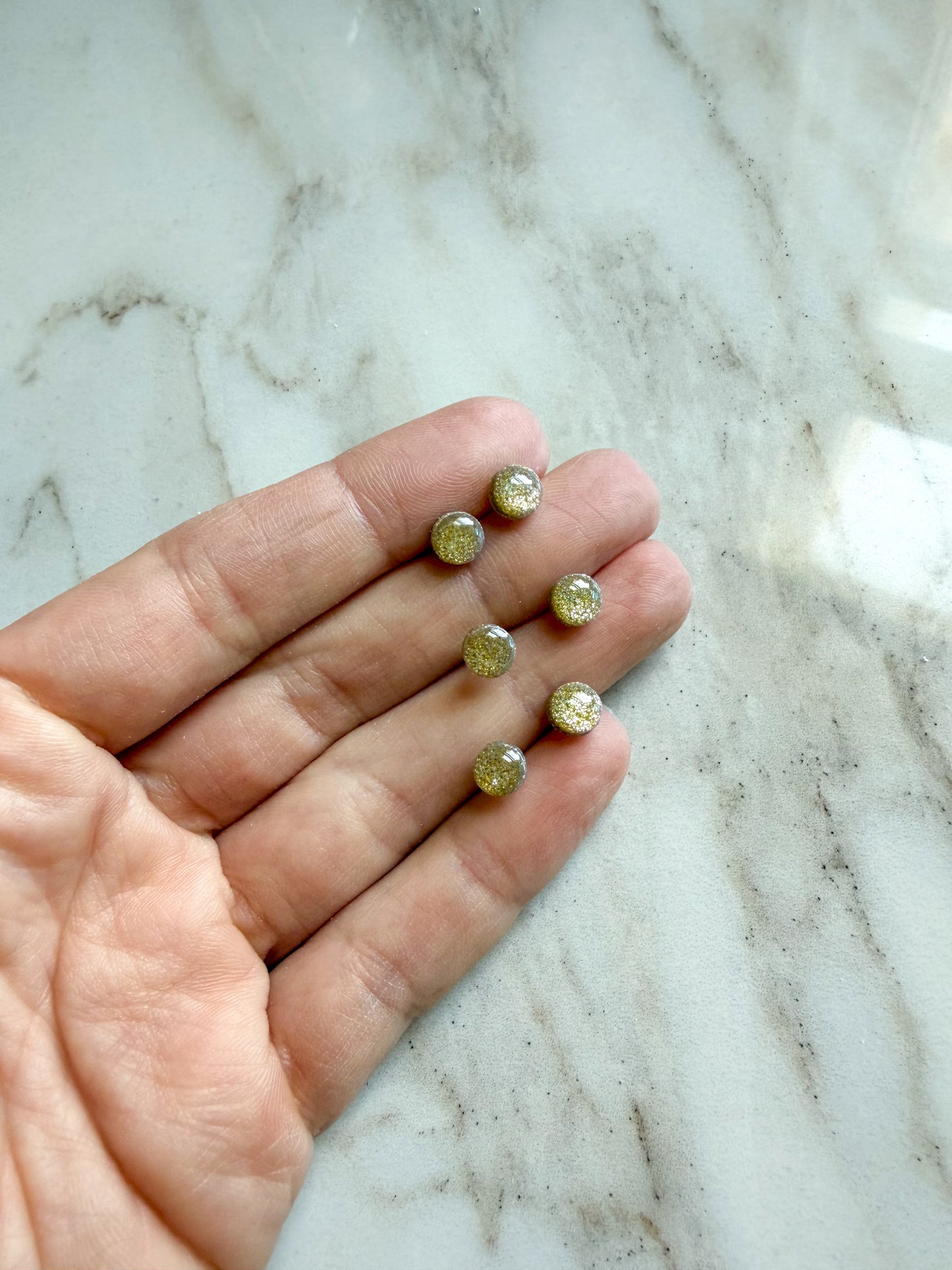 Six small round gold-toned earrings with textured glitter interiors are displayed on a person's fingers against a white and gray marble background.