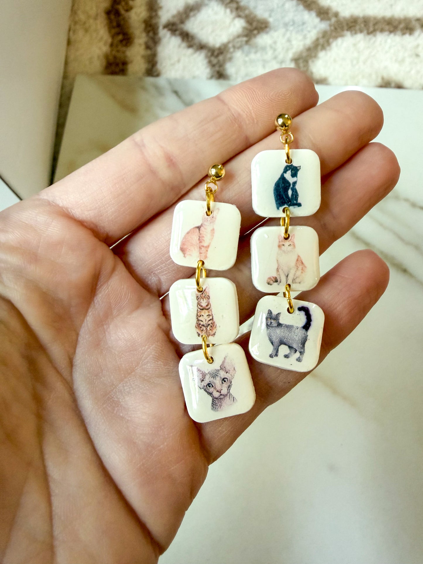 A pair of earrings held in a hand. They each have 3 squares with a cat in the centers.