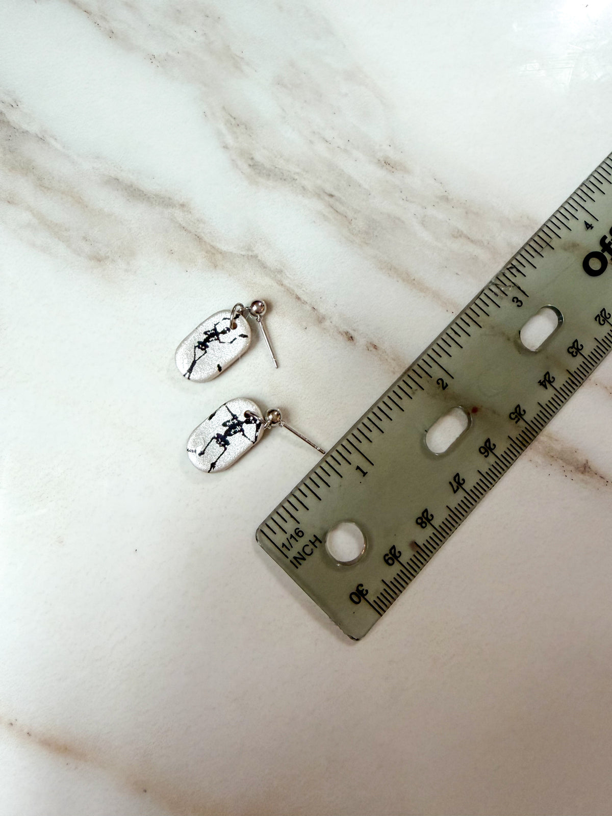A pair of silver-toned dangle earrings with white oval charms printed with black skeleton art, shown next to a ruler on a light marble surface. About 1 inch in length. 