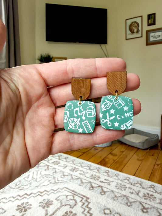 A hand holding a pair of earrings against a living room background. They are green to resemble old chalkboards, with white school-themed scribble designs. The posts are wood. 