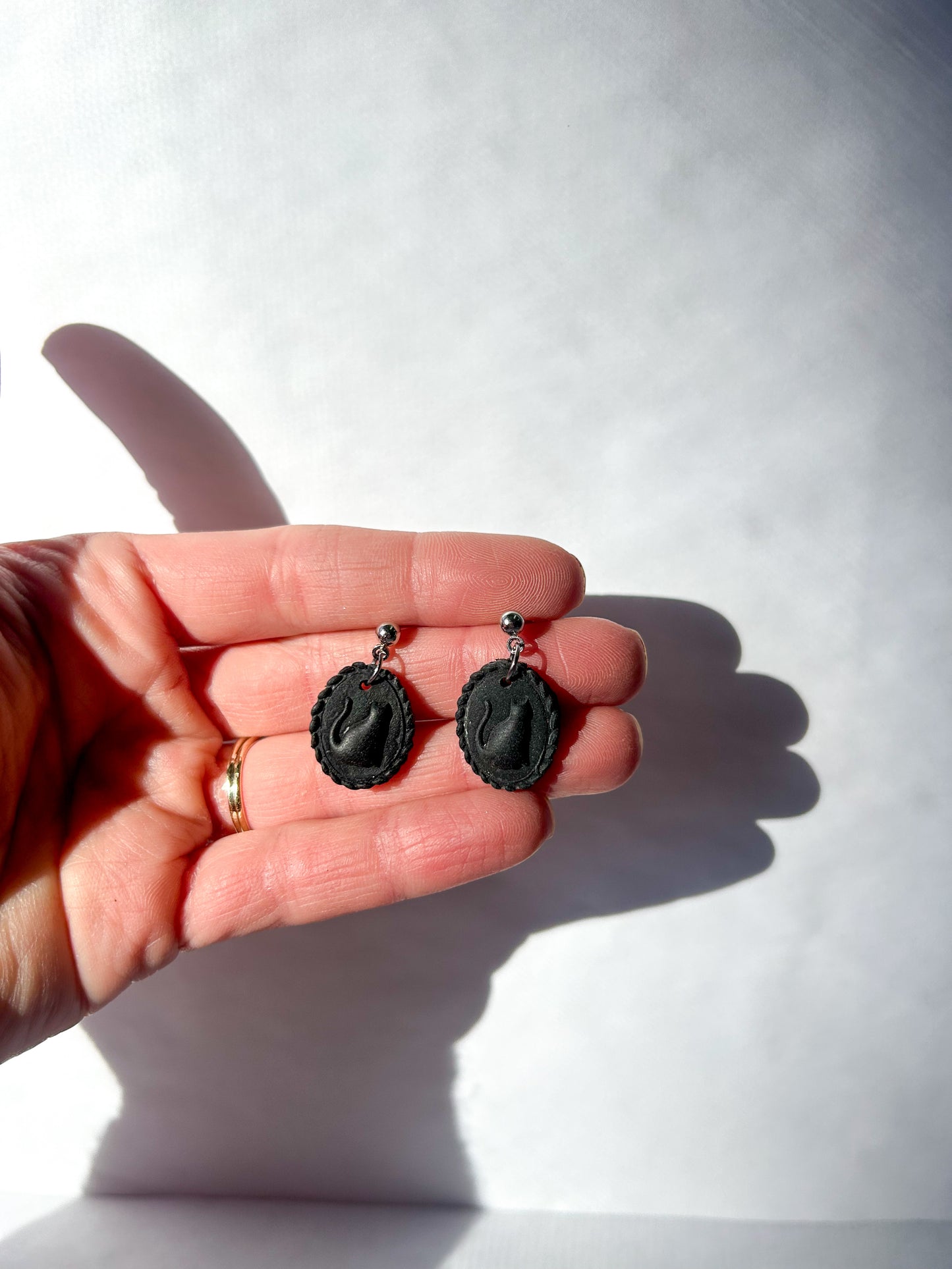 Black earrings held in a hand against a white background. The earrings resemble a scalloped frame with a raised cat silhouette in the center.
