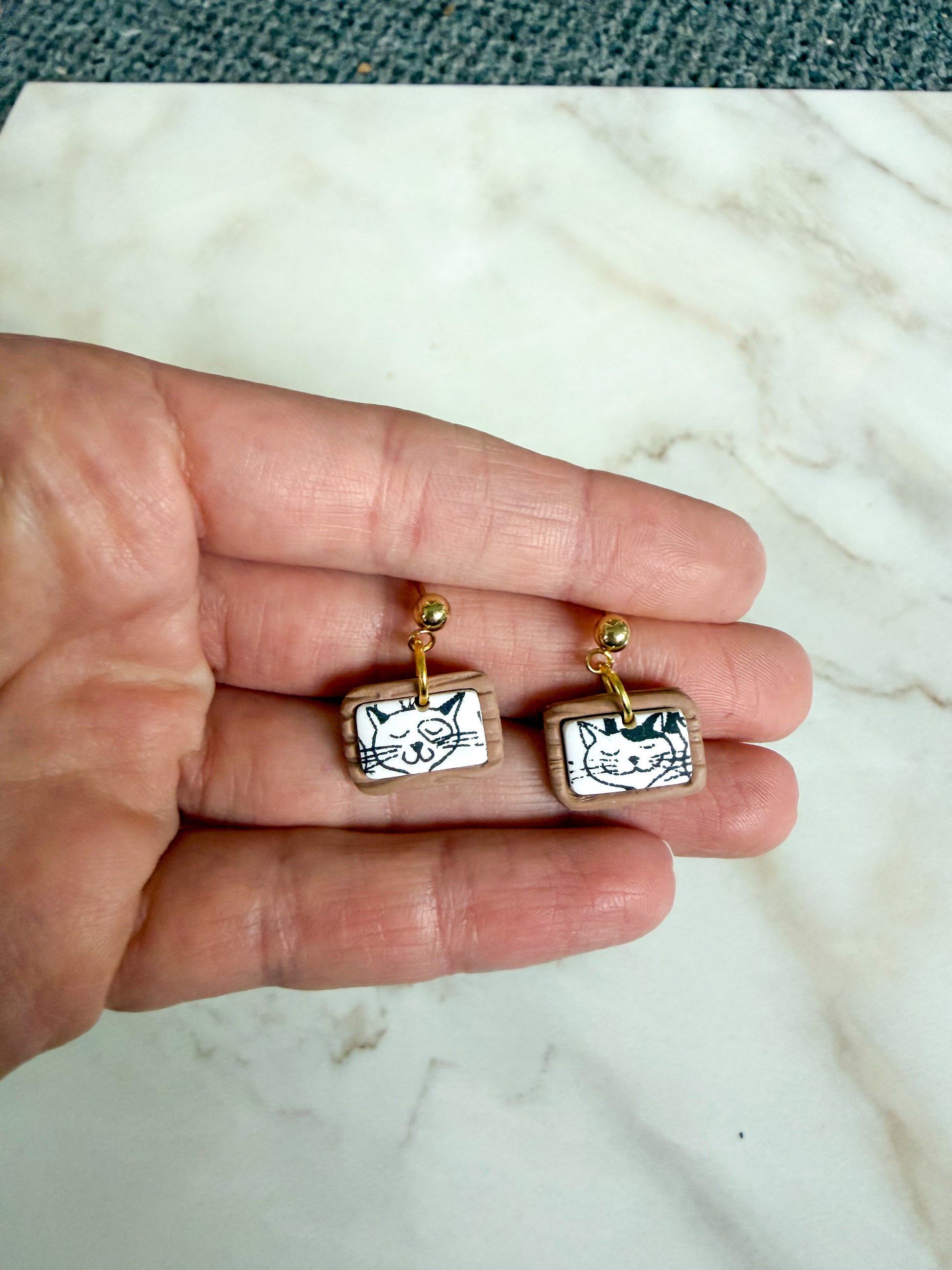 Mini dangles that look like a picture of a cartoon cat in a wooden frame, held in a hand. 
