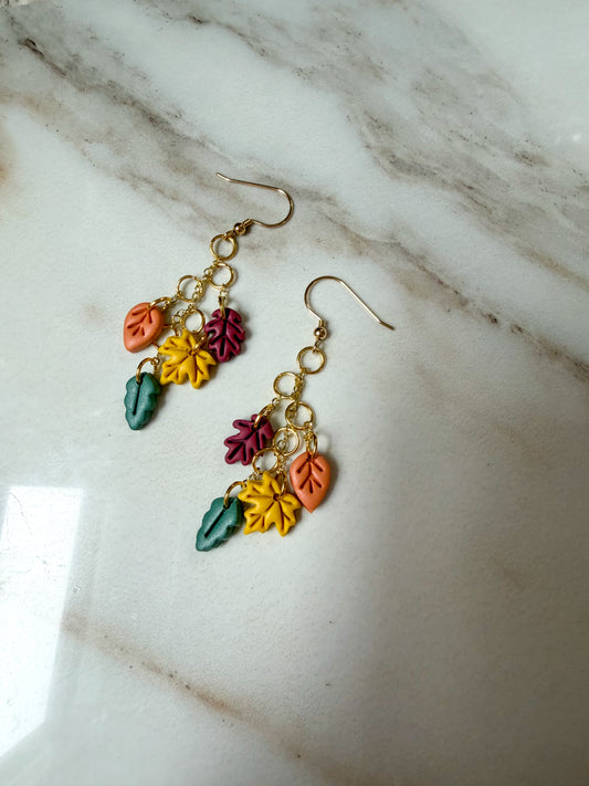 A pair of gold-tone dangle earrings with colorful autumn leaf charms in orange, yellow, green, and burgundy, displayed on a white marble surface.