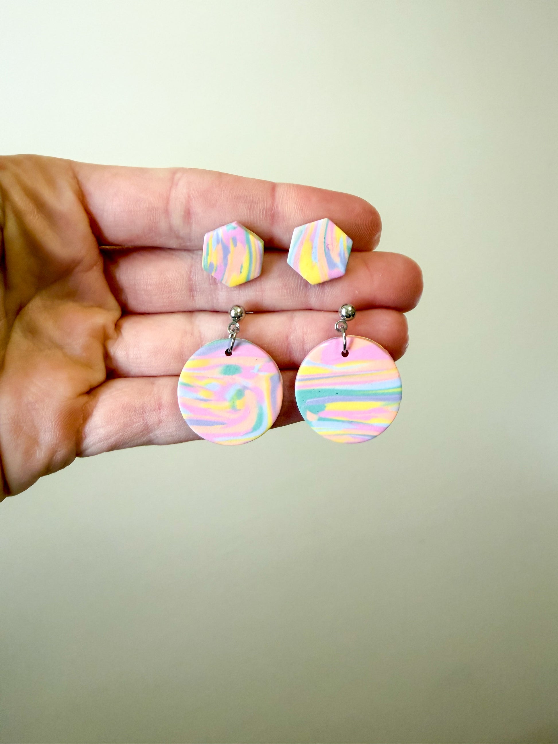 A hand holding a pair of pastel marbled earrings, featuring small hexagonal studs and larger round drop pendants in soft pink, yellow, blue, and white swirls against a plain light background.