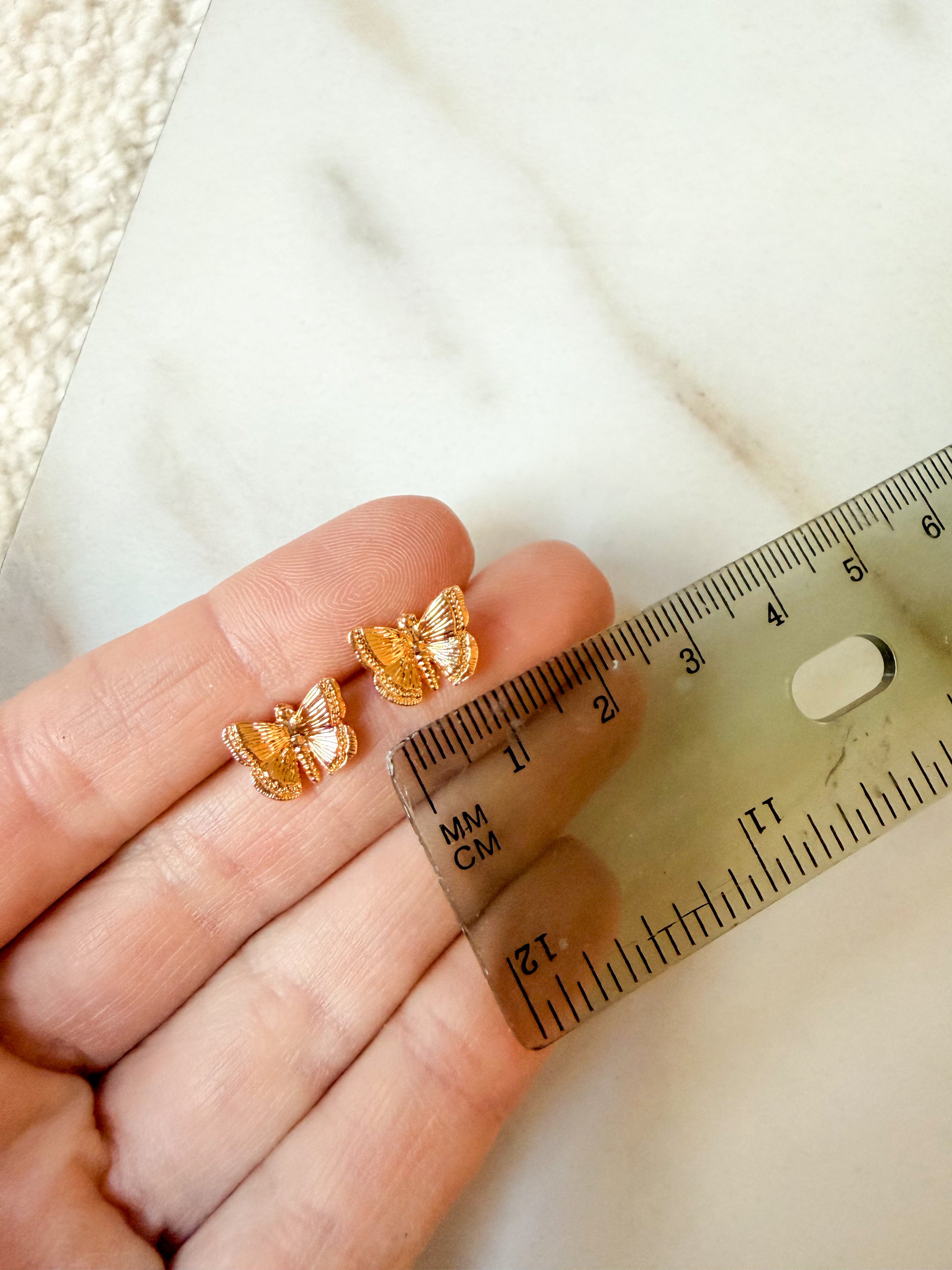 A close-up of a hand holding a pair of small gold butterfly-shaped stud earrings next to a clear ruler for scale, with a light marble surface in the background. About one centimeter in length. 