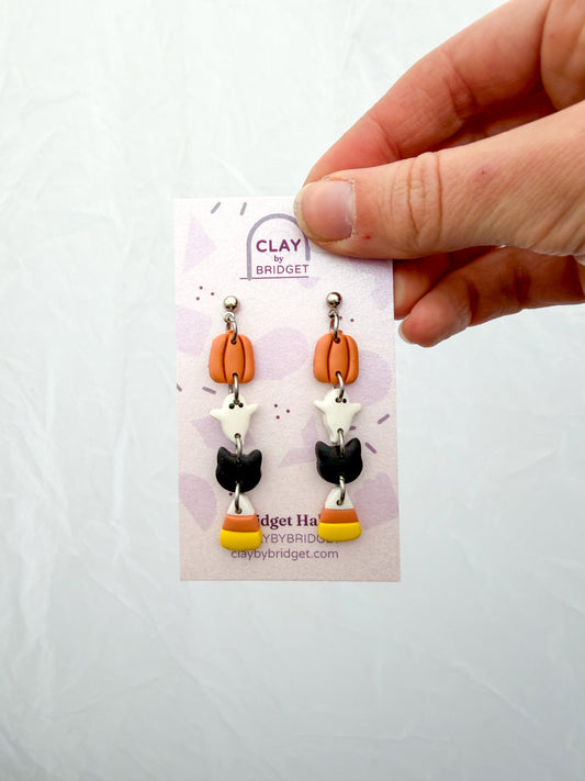 Hand holding a pair of earrings on a card. They feature tiny charms stacked and connected with jump rings. A pumpkin, ghost, black cat, and candy corn. 