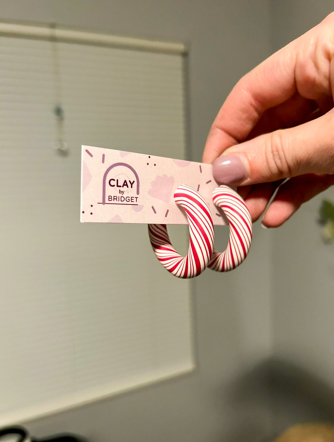 Hand holding a card with 'CLAY by Bridget' and two red and white peppermint striped clay earrings.
