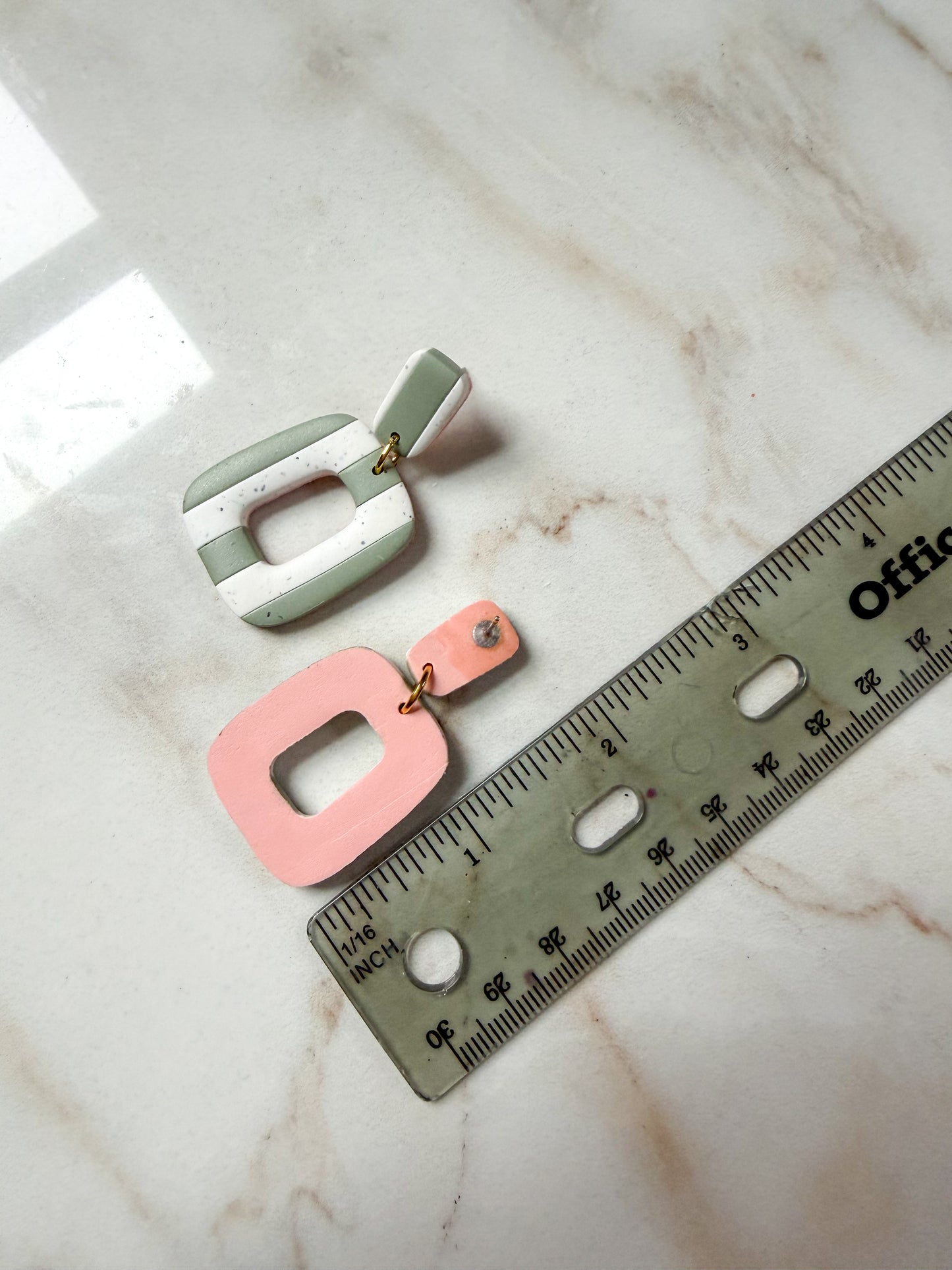 Two earrings, one green and one pink, on a marble surface with a ruler for scale.