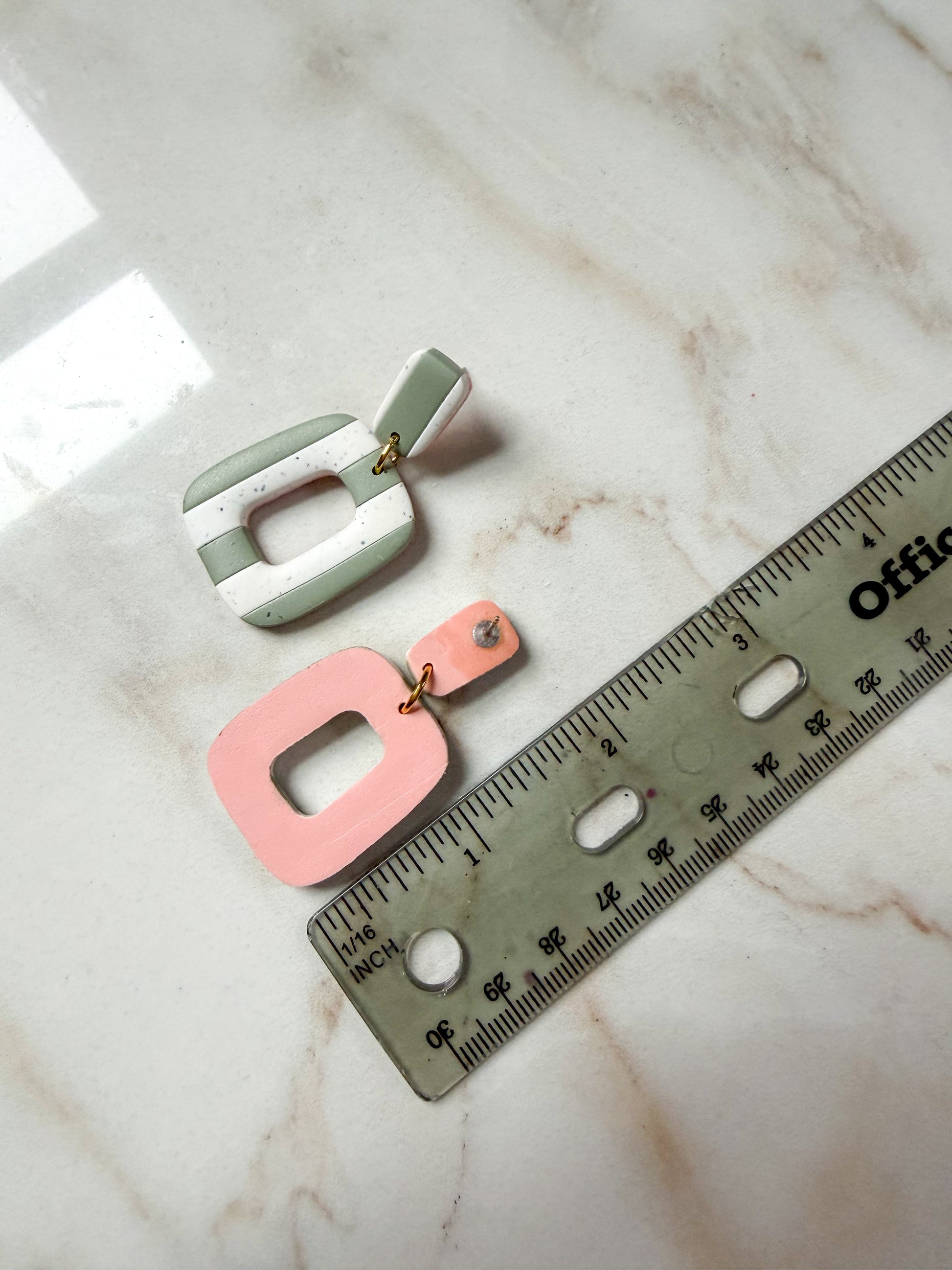 Two earrings, one green and one pink, on a marble surface with a ruler for scale.