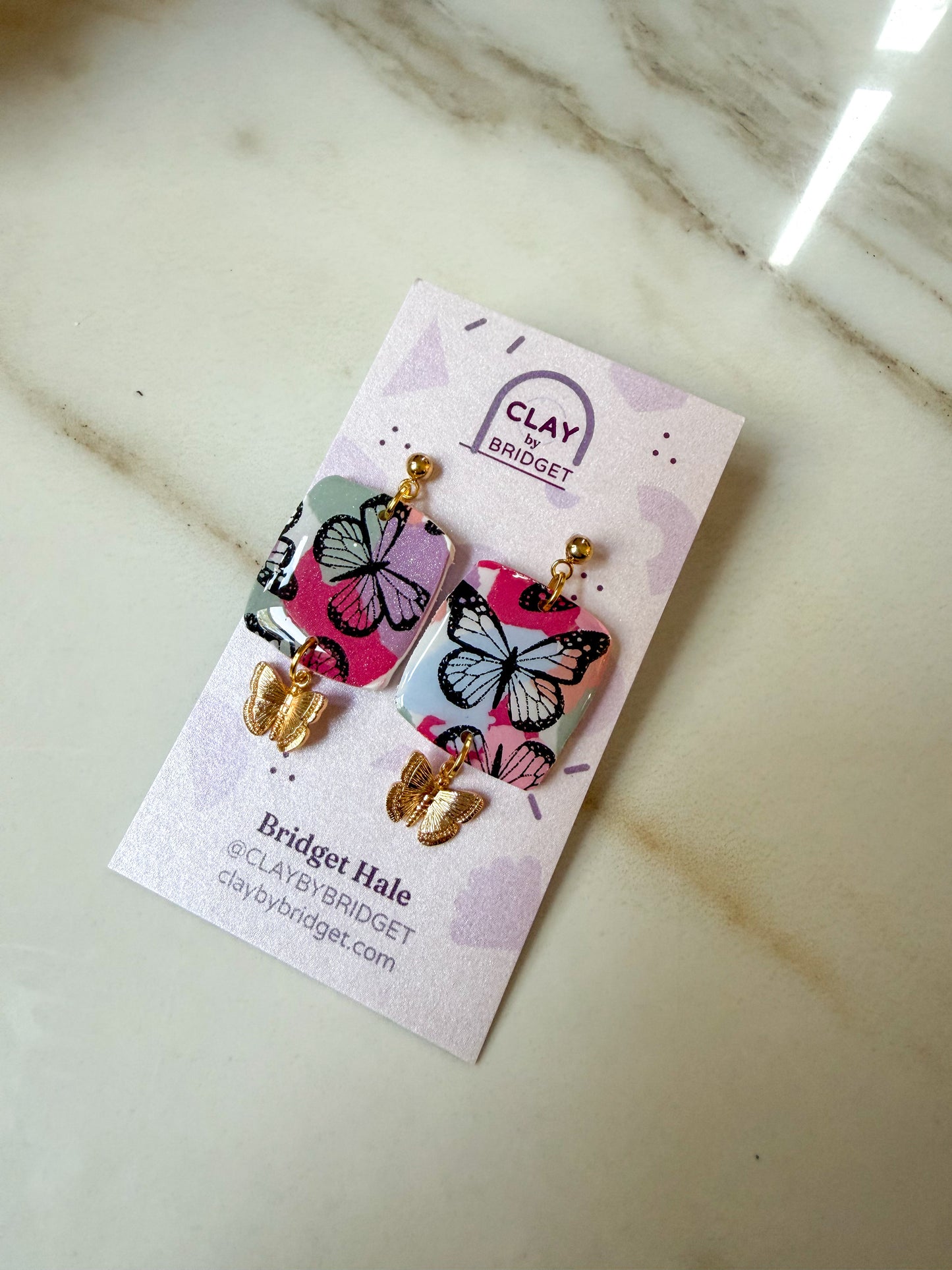 A pair of butterfly-themed drop earrings on a branded display card, photographed on a light marble surface. The earrings have pink and blue butterfly charms with black outlines and gold-tone hooks and beads.