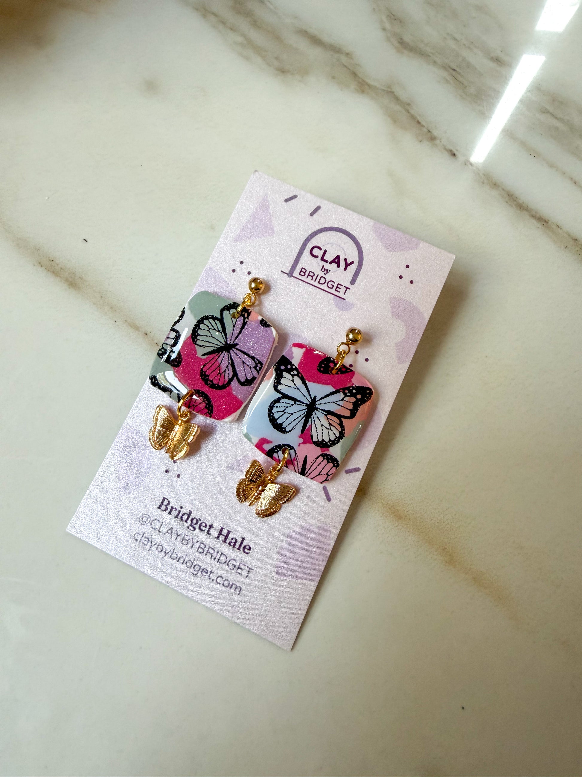 A pair of butterfly-themed drop earrings on a branded display card, photographed on a light marble surface. The earrings have pink and blue butterfly charms with black outlines and gold-tone hooks and beads.