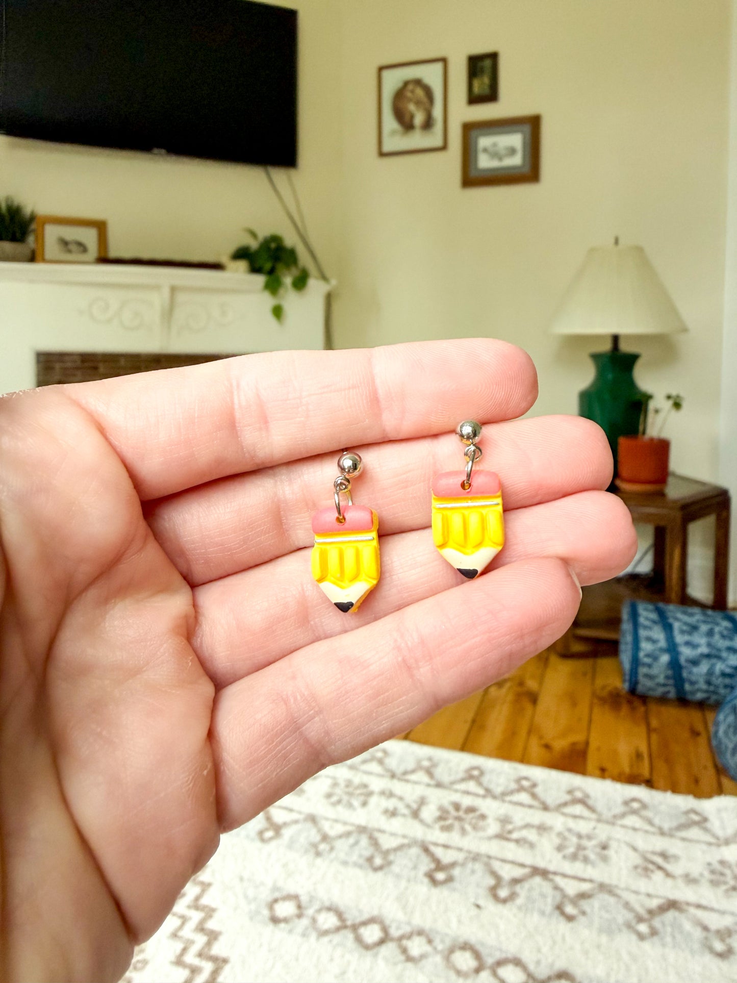 A hand holding a pair of yellow and pink pencil-shaped earrings with silver posts, set against a blurred living room background.