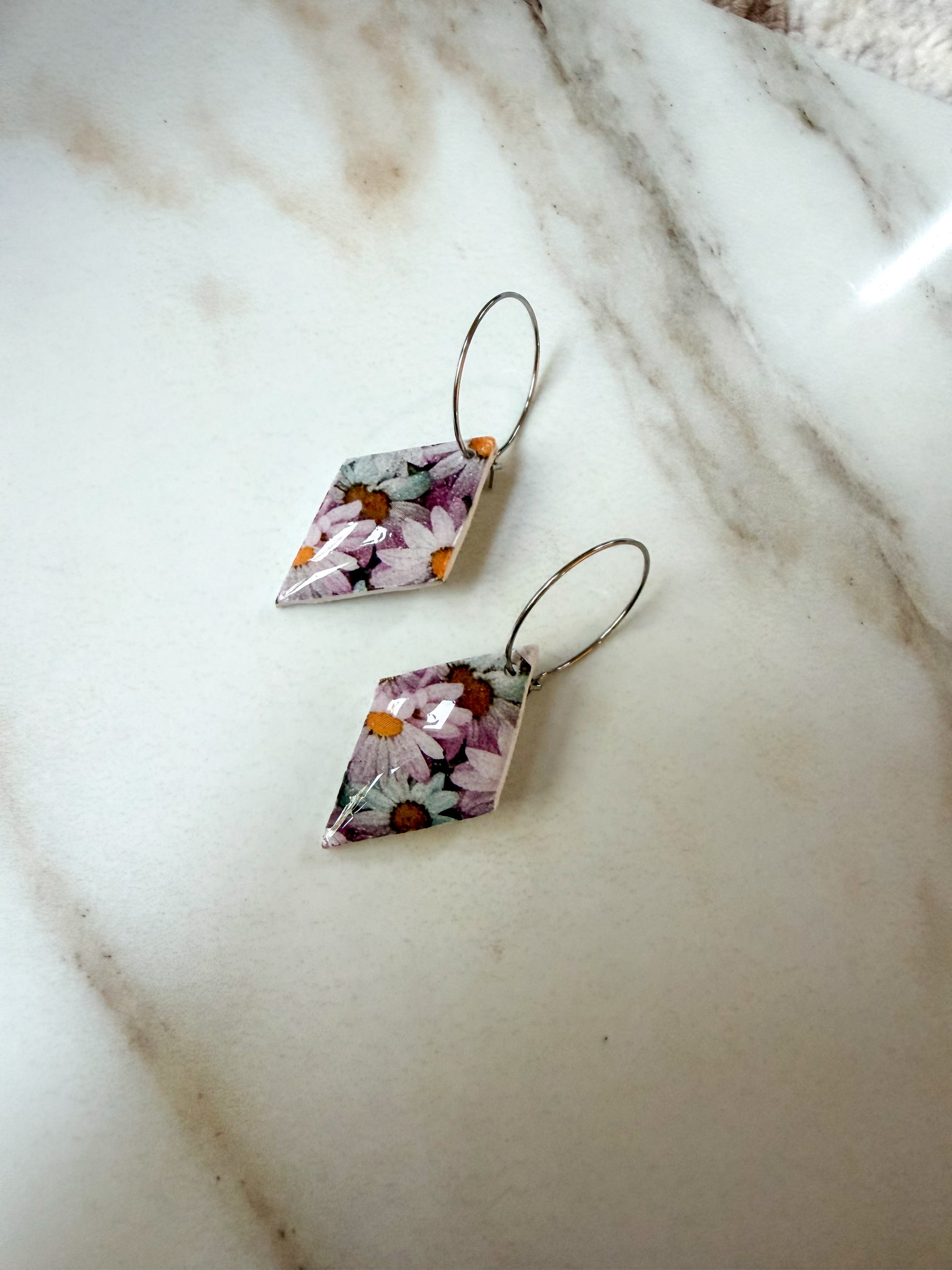 A pair of dangling earrings with irregular diamond-shaped floral resin pendants and thin silver-tone wire hooks, resting on a white marble surface.