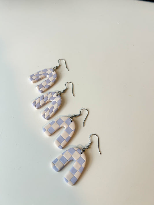 Two pairs of arch shaped earrings laying on a flat surface. The pair furthest from the camera has a swirled periwinkle and cream checker pattern. The pair closest has the same colors, but the lines of the checker pattern are straight. 