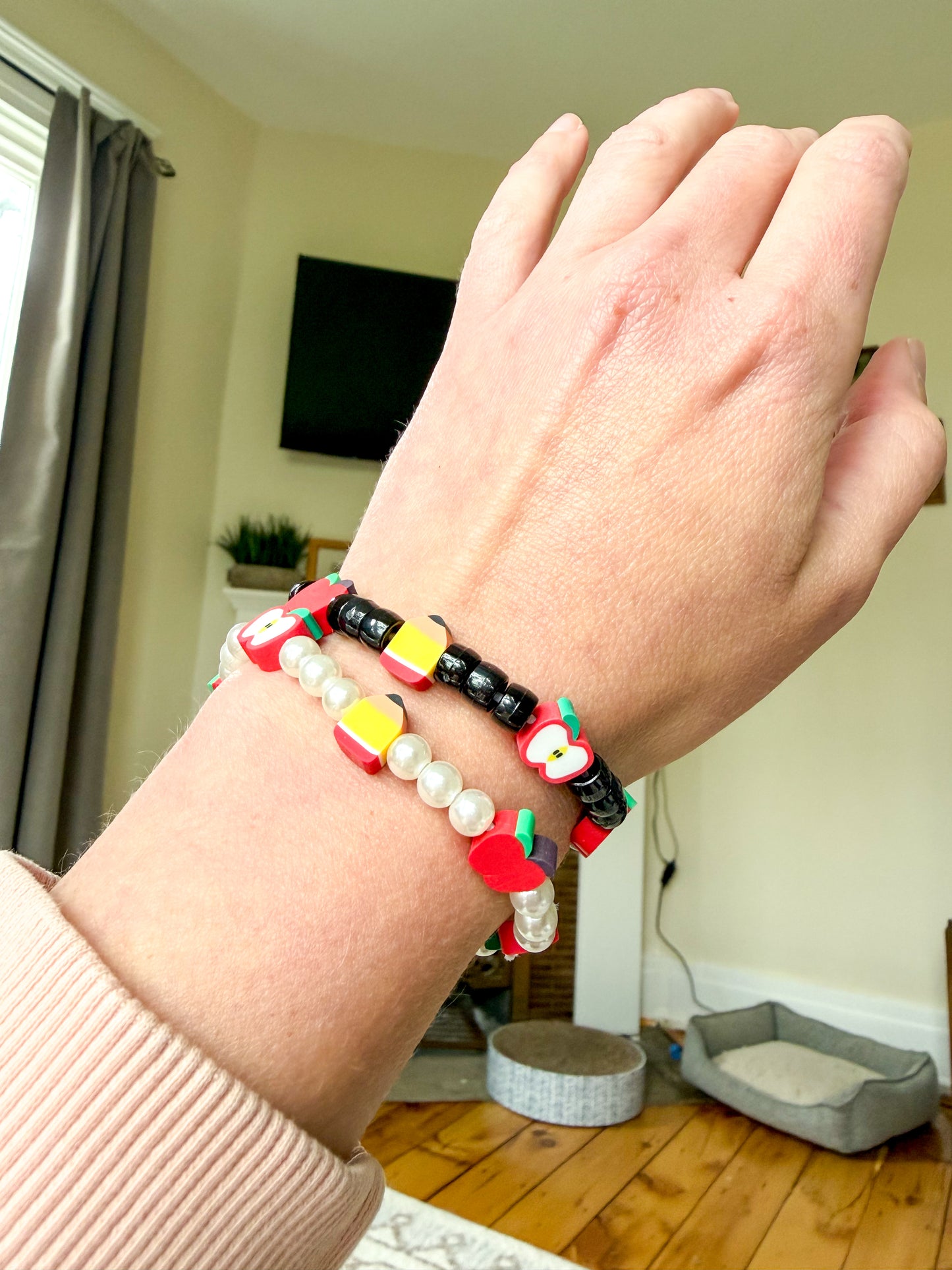 A close-up of a person's wrist wearing two colorful beaded bracelets in a home setting with a wooden floor and pet bed visible in the background.