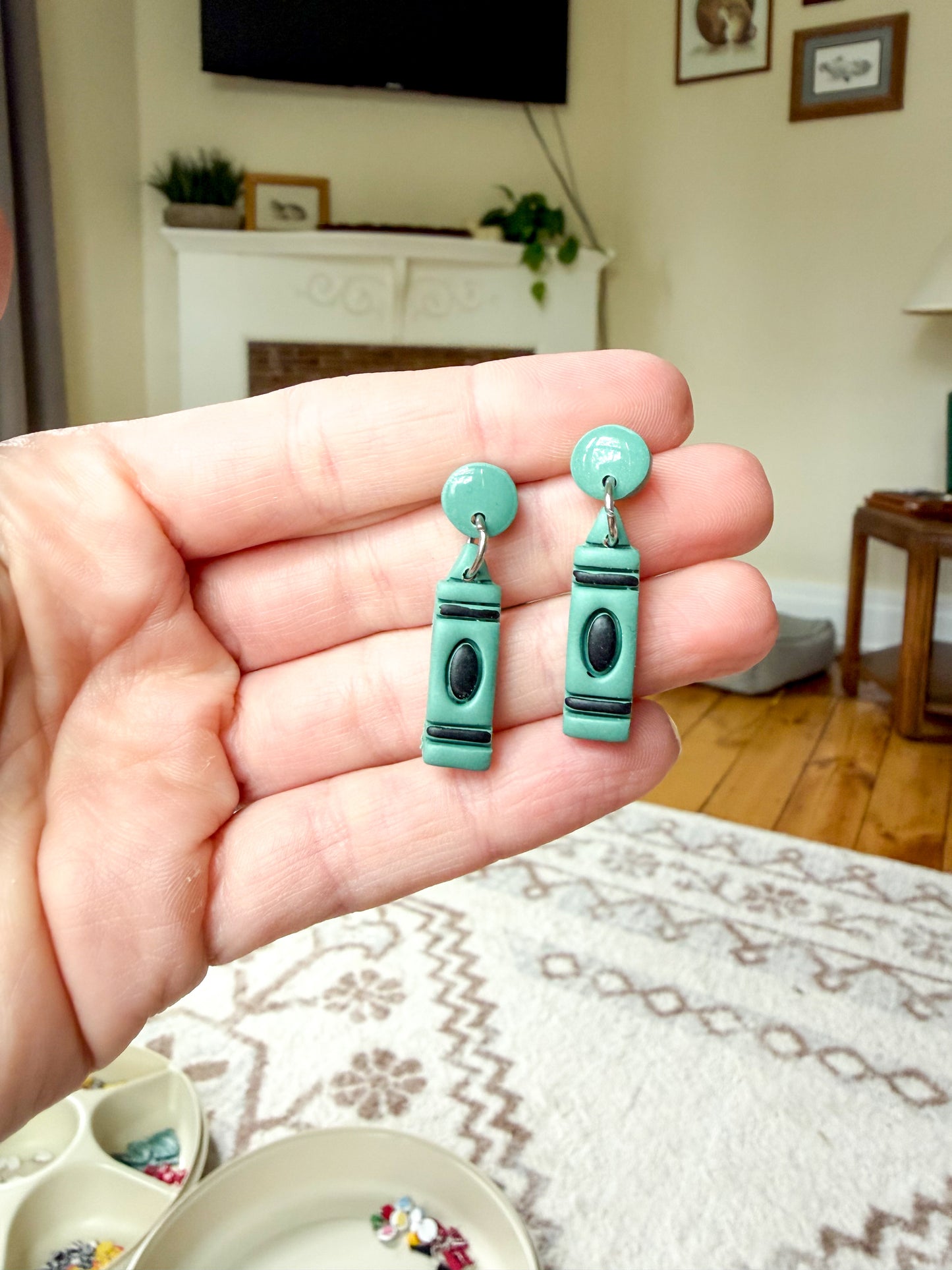 A hand holds a pair of green polymer clay earrings shaped like crayons, with black and silver details.