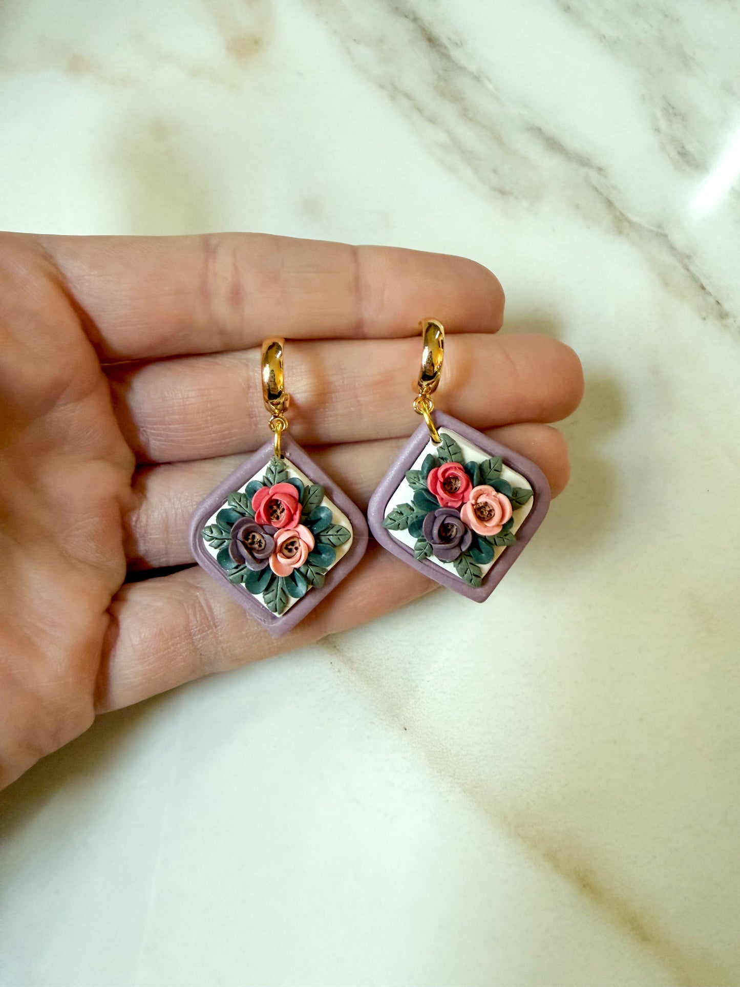 A pair of handmade square dangle earrings with gold hooks and lavender-purple frames, featuring a 3D floral design with pink, red, and purple roses and green leaves, held in a hand over a white marble background.