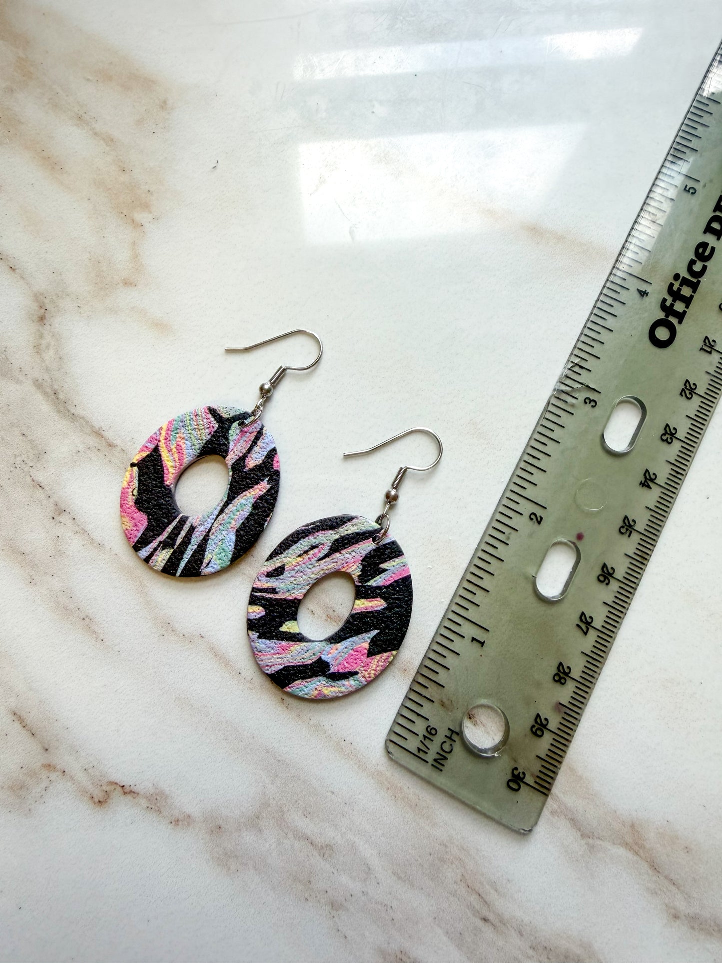 Colorful oval dangles with a center hole cut out. The pattern is black with colorful swirls. They are lying on a marble surface next to a ruler, and are about 2 inches in length. 