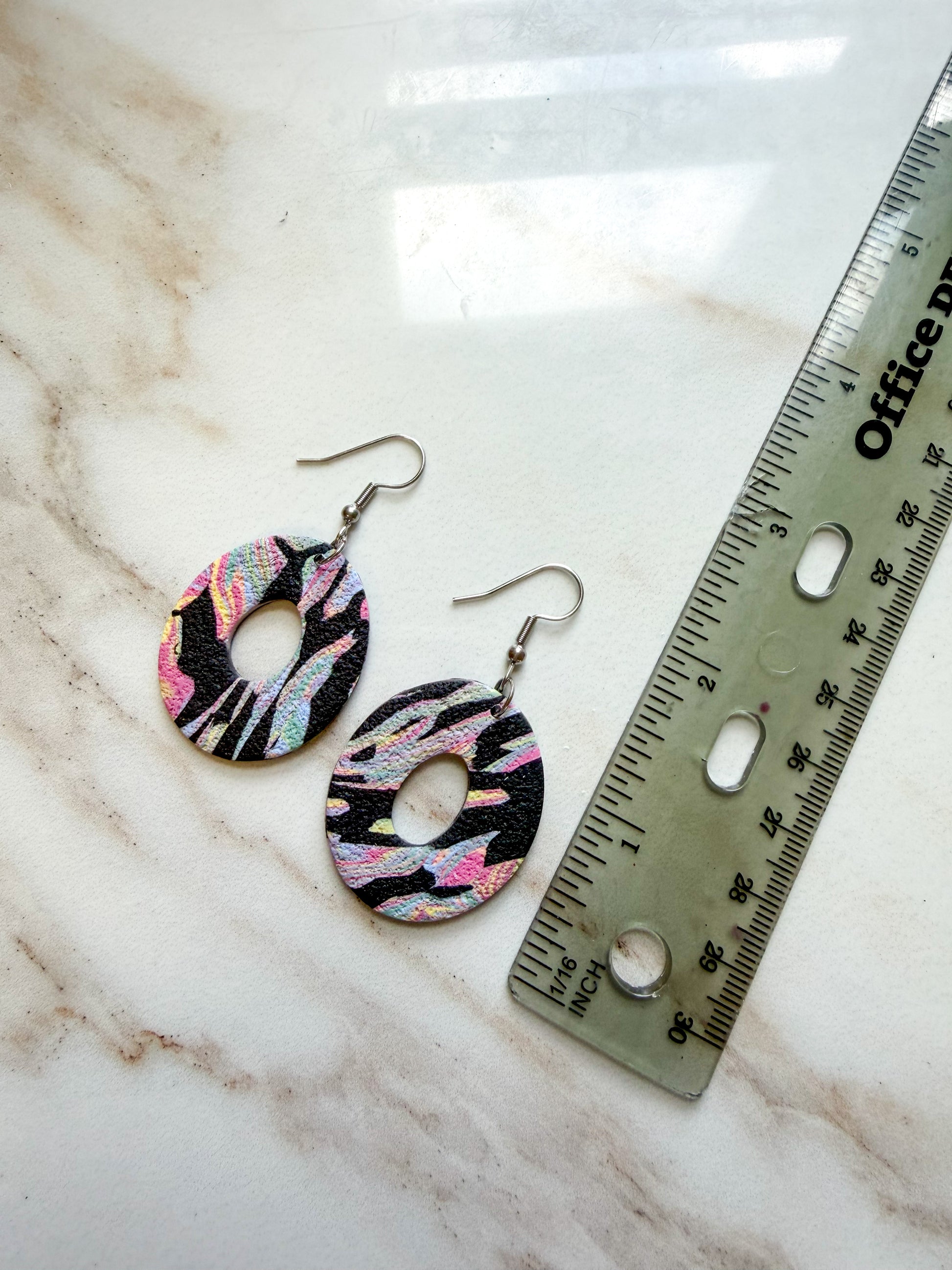 Colorful oval dangles with a center hole cut out. The pattern is black with colorful swirls. They are lying on a marble surface next to a ruler, and are about 2 inches in length. 