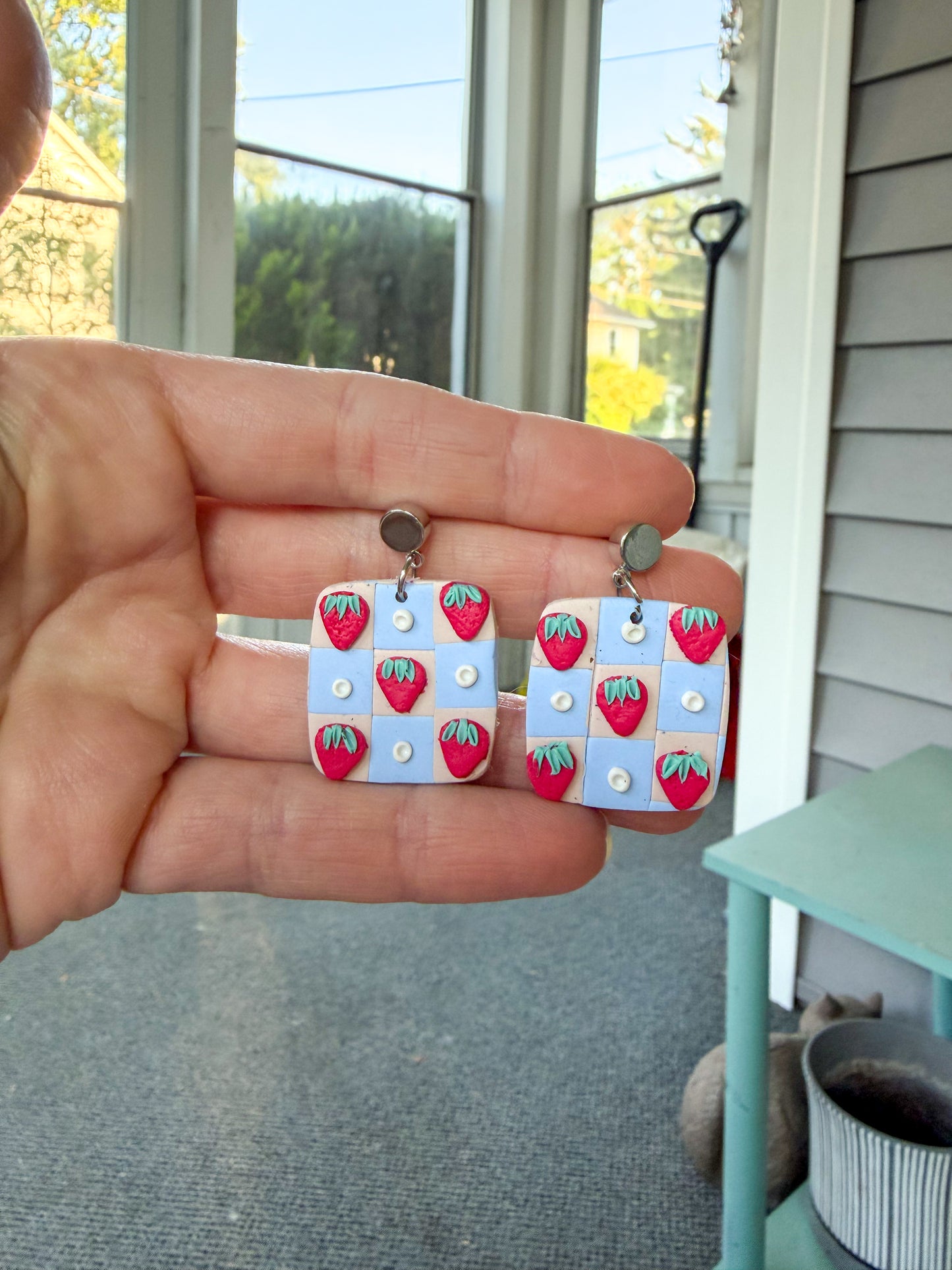 Hand holding a pair of earrings with strawberry design in front of a window.