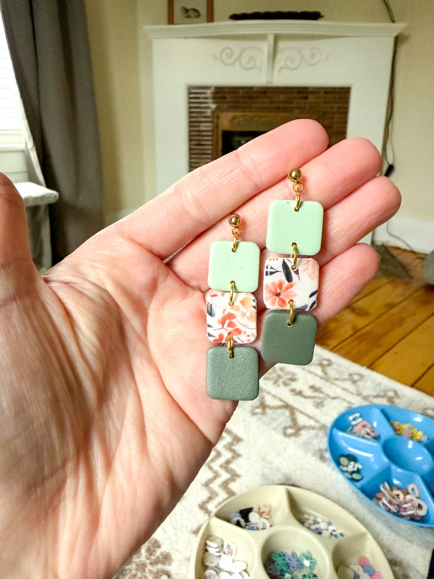 Hand holding a pair of colorful square-shaped earrings with floral designs.