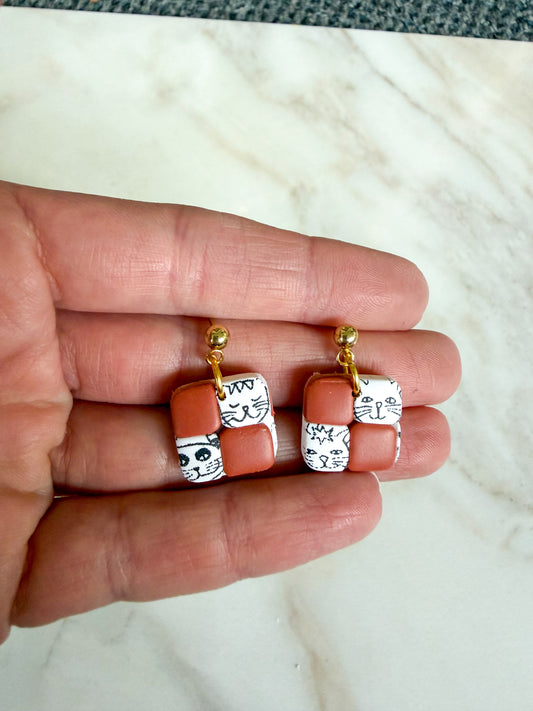 Pair of earrings with cat designs held between fingers on a marble surface