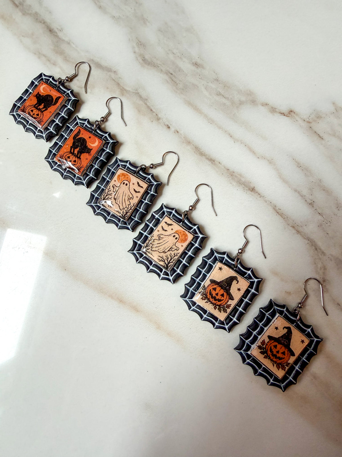 Six Halloween-themed dangle earrings arranged diagonally on a white marble surface. Each earring has a silver fishhook wire and a shield-shaped charm with a black spiderweb border, orange center, and a different spooky design: a cat, a pumpkin, a ghost, and a jack-o'-lantern.