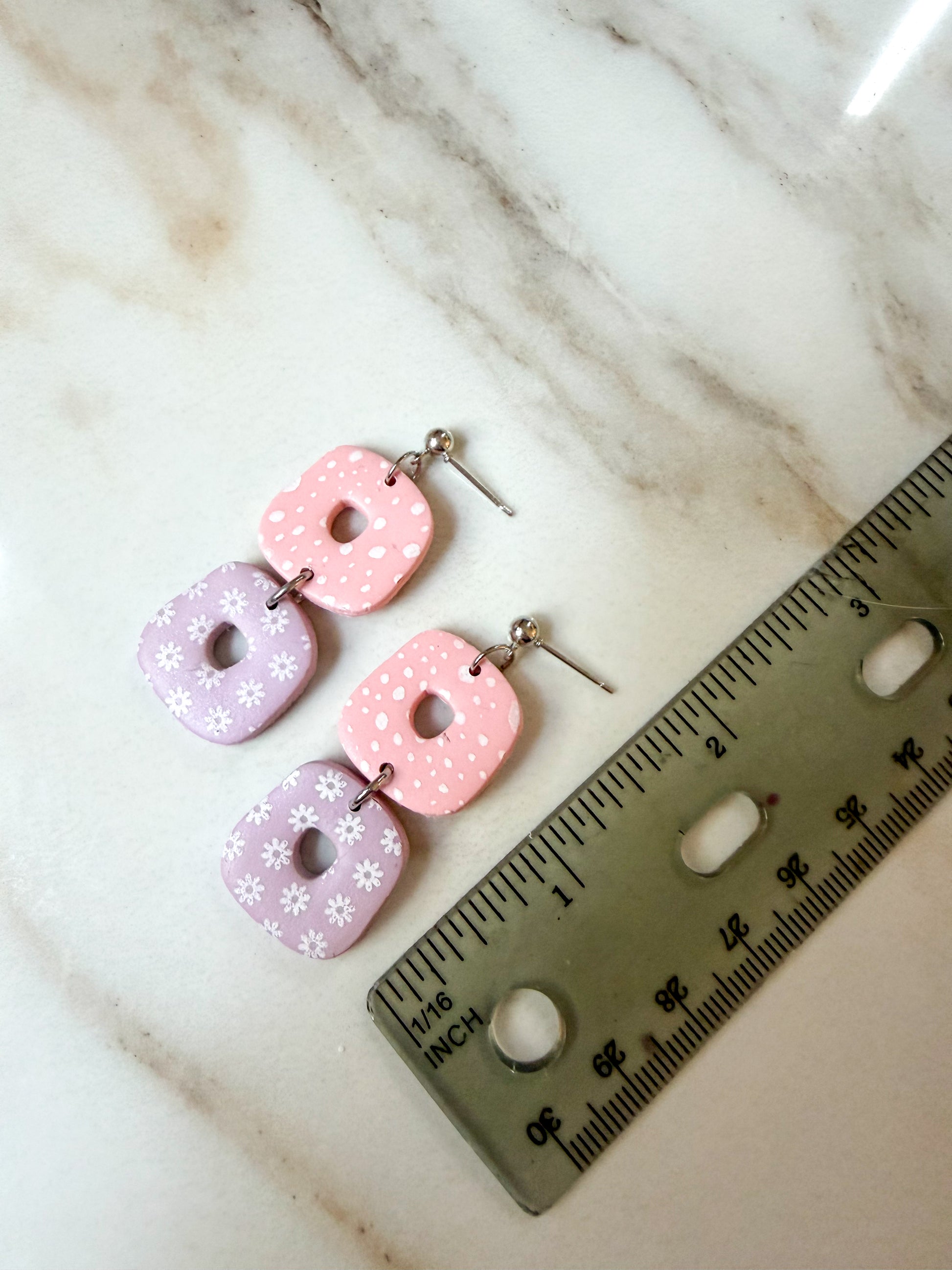 A pair of handmade dangle earrings with pink and lavender donut-shaped charms decorated with white polka dots and floral prints, displayed next to a ruler on a white marble surface. 2 inches in length. 