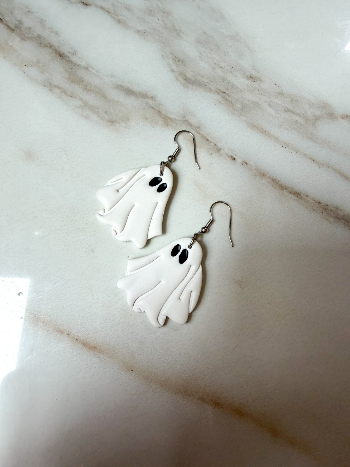 A pair of white ghost-shaped dangle earrings with black eyes and silver fishhook wires, displayed on a white and gray marble surface.