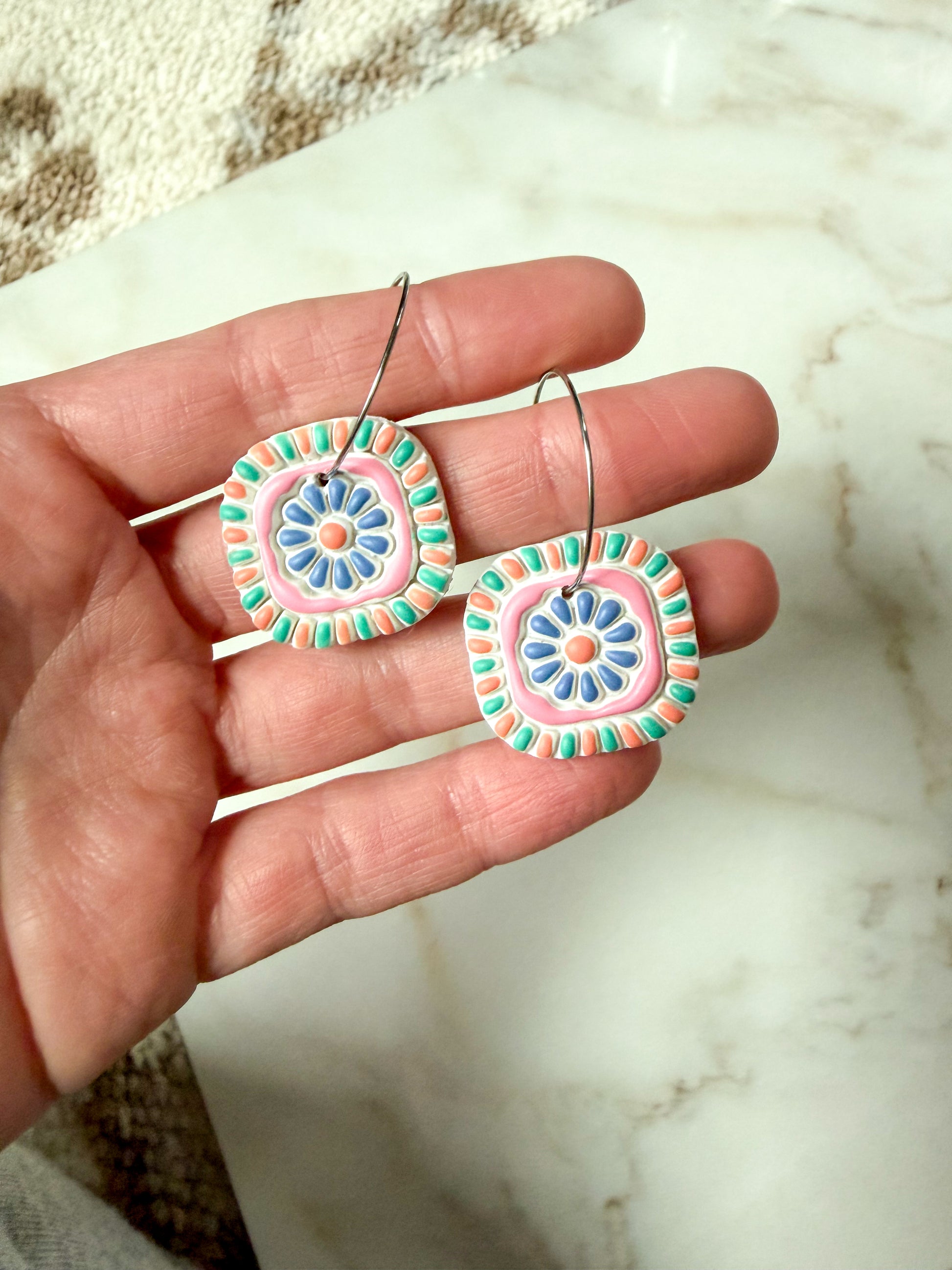 Hand holding a pair of colorful circular earrings on a marble surface