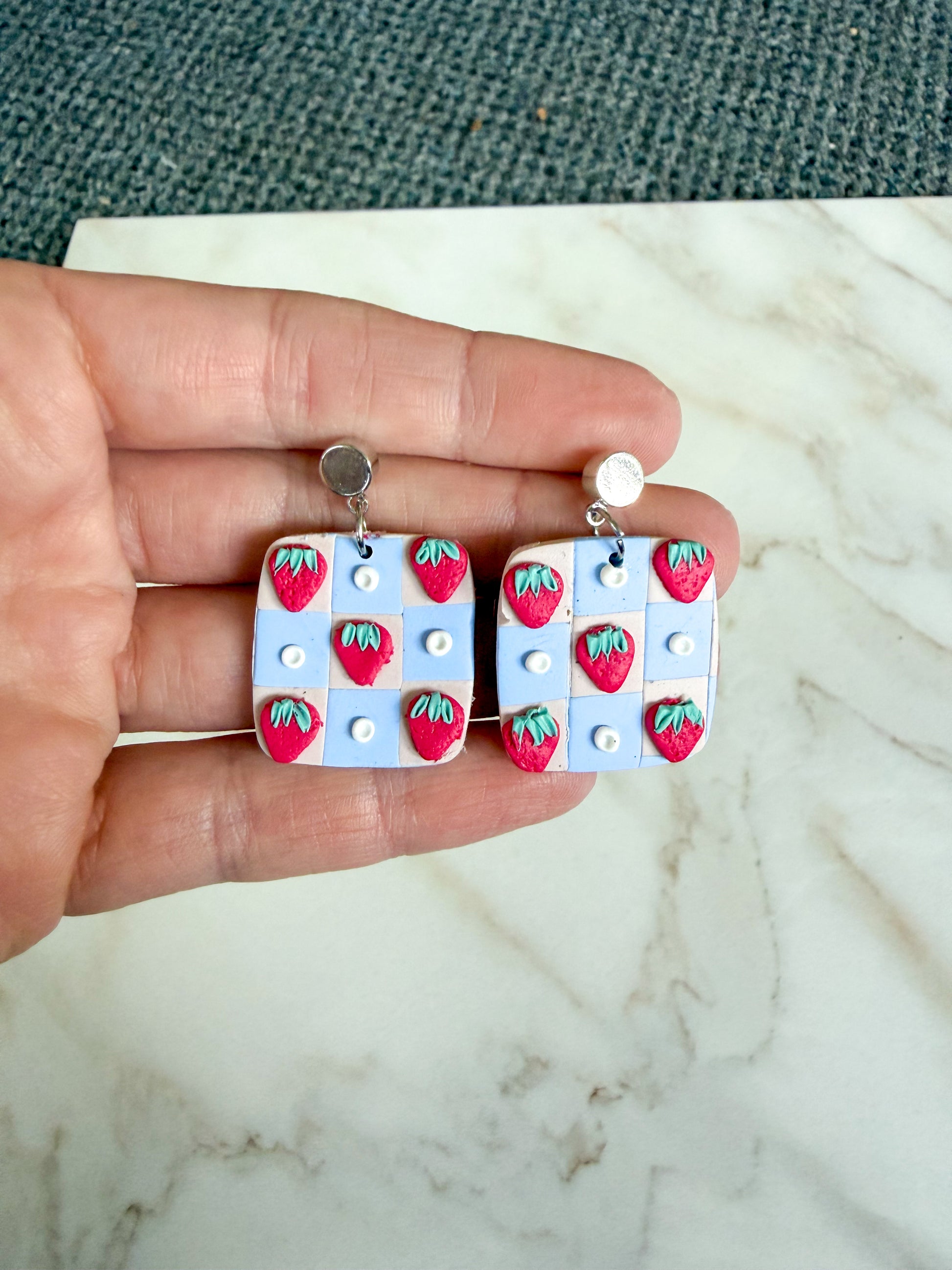 Hand holding a pair of earrings with strawberry design on a marble surface