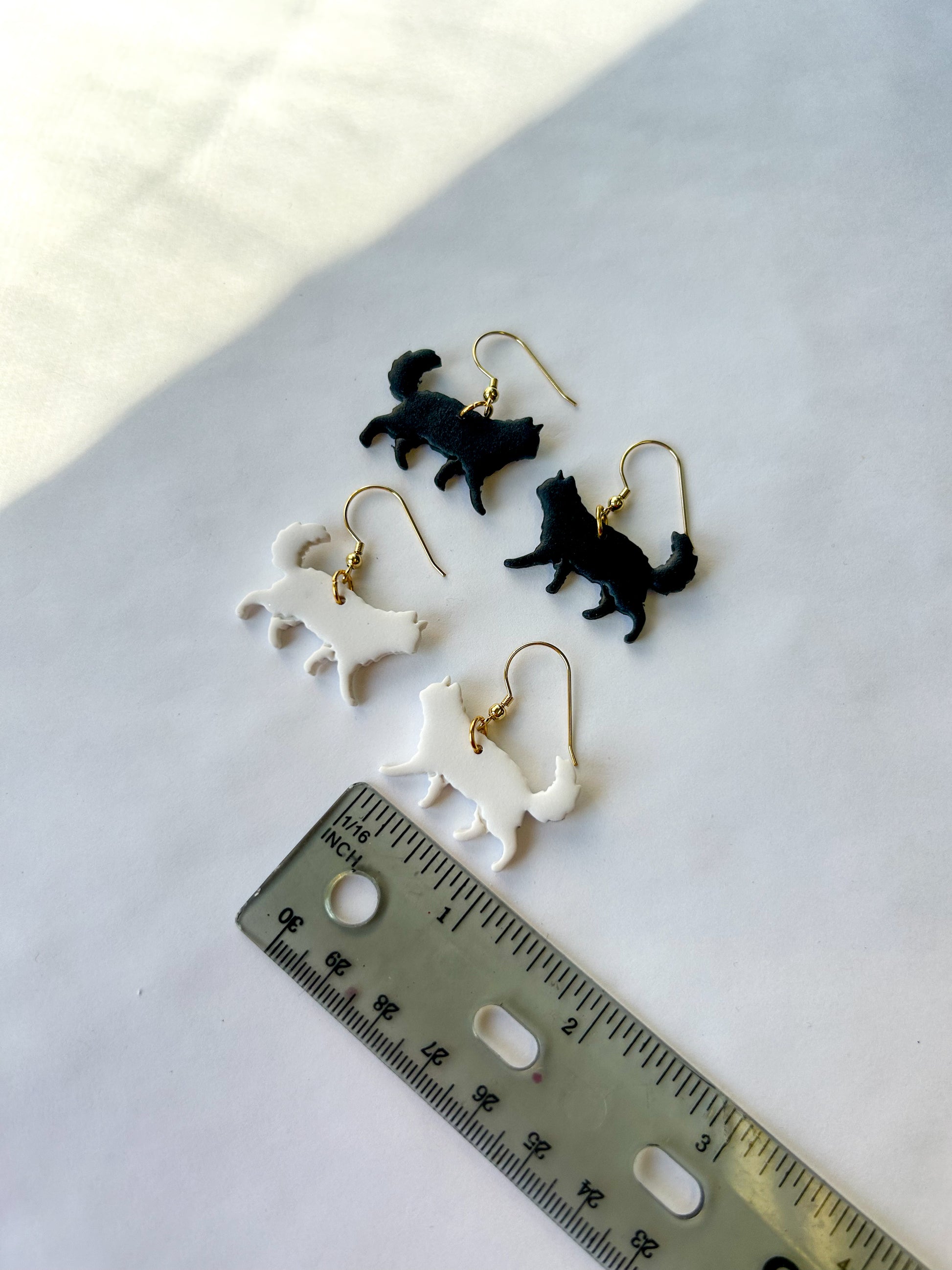 Cat-shaped earrings on a white surface with a ruler for scale. One pair is black, and the other pair is white. Long-haired cats