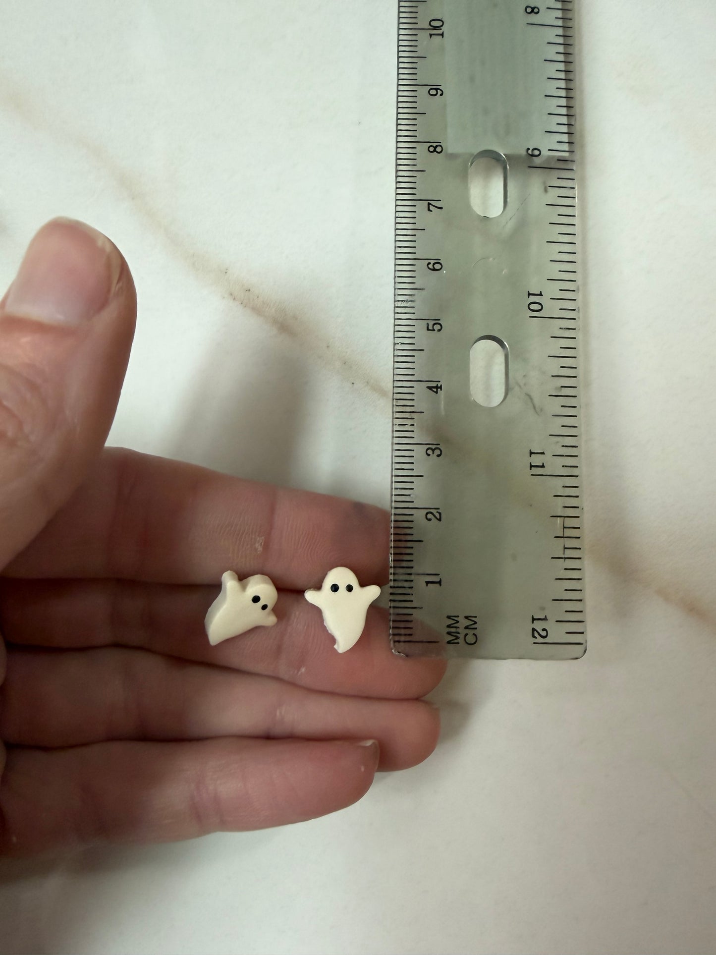 Two small white ghost-shaped items resting on a person's fingers next to a ruler for scale, photographed on a light marble-like surface. About 1cm long. 