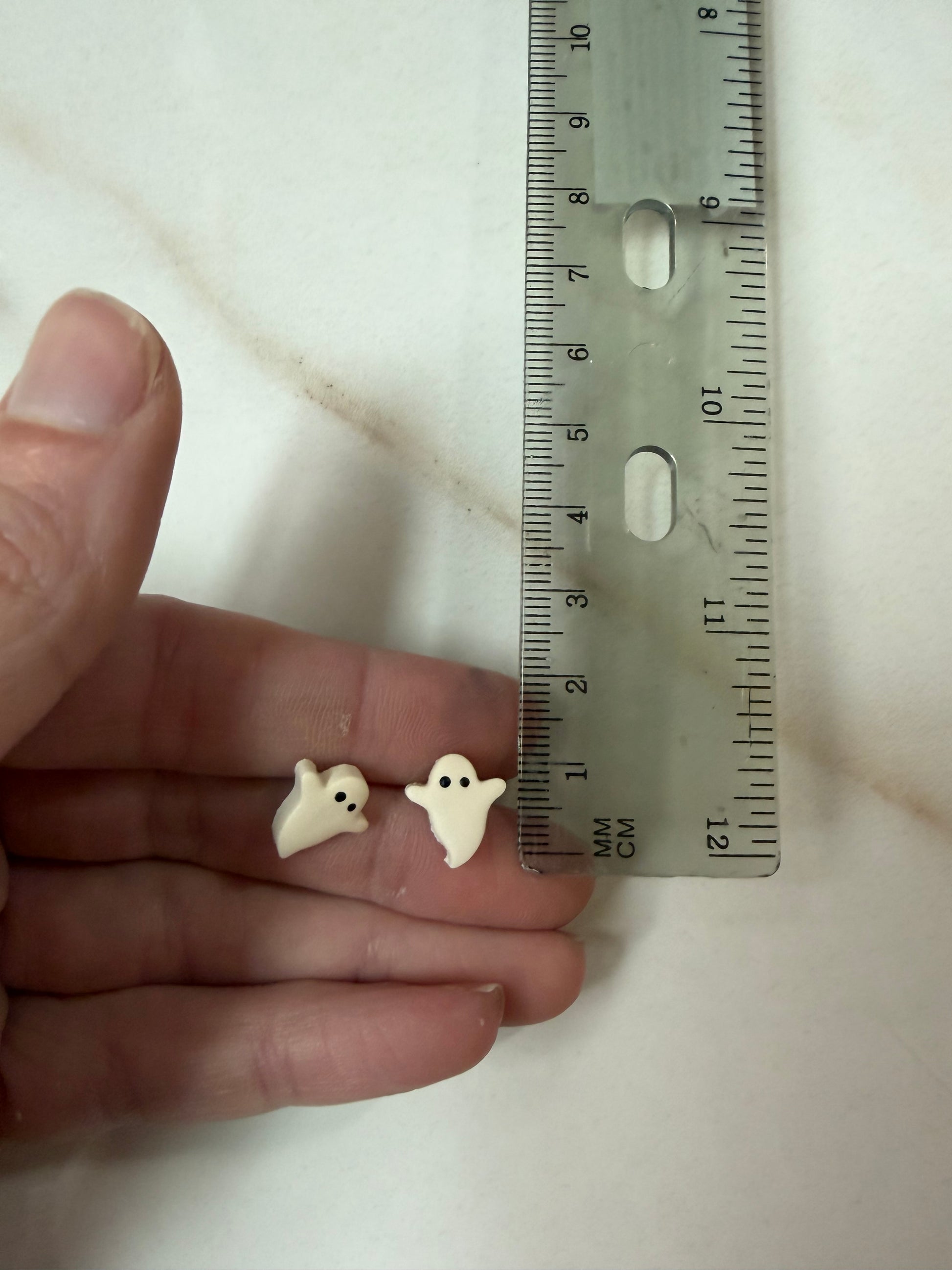 Two small white ghost-shaped items resting on a person's fingers next to a ruler for scale, photographed on a light marble-like surface. About 1cm long. 