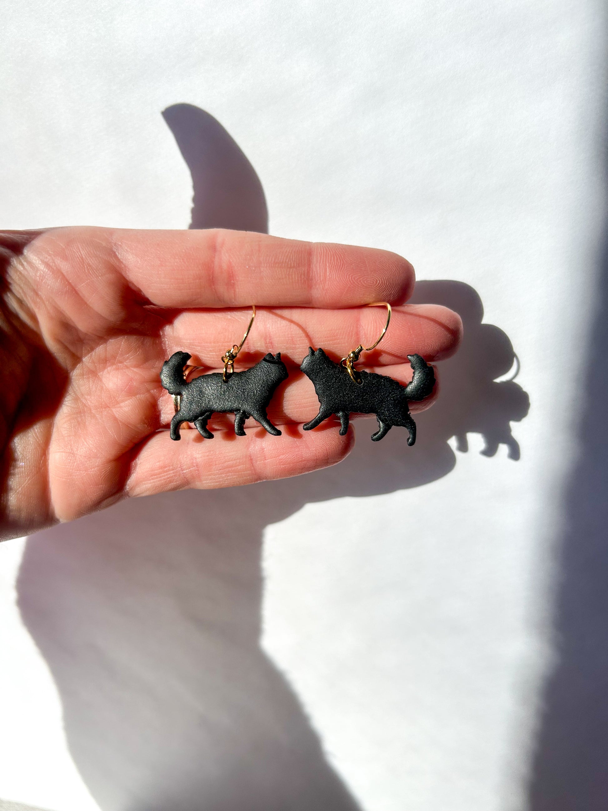 Hand holding long haired black cat-shaped earrings against a white background
