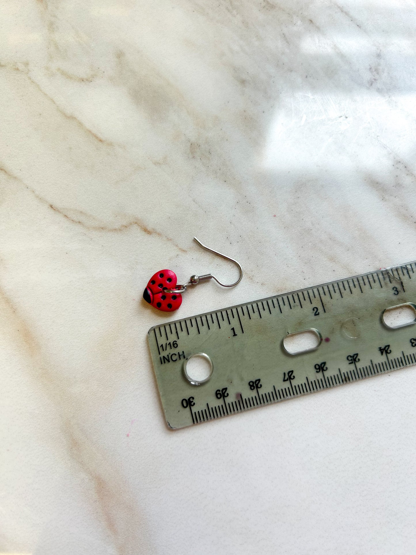 Heart shaped earrings that are painted to look like ladybugs, laying next to a ruler to demonstrate that they are approximately 1 inch in length ￼