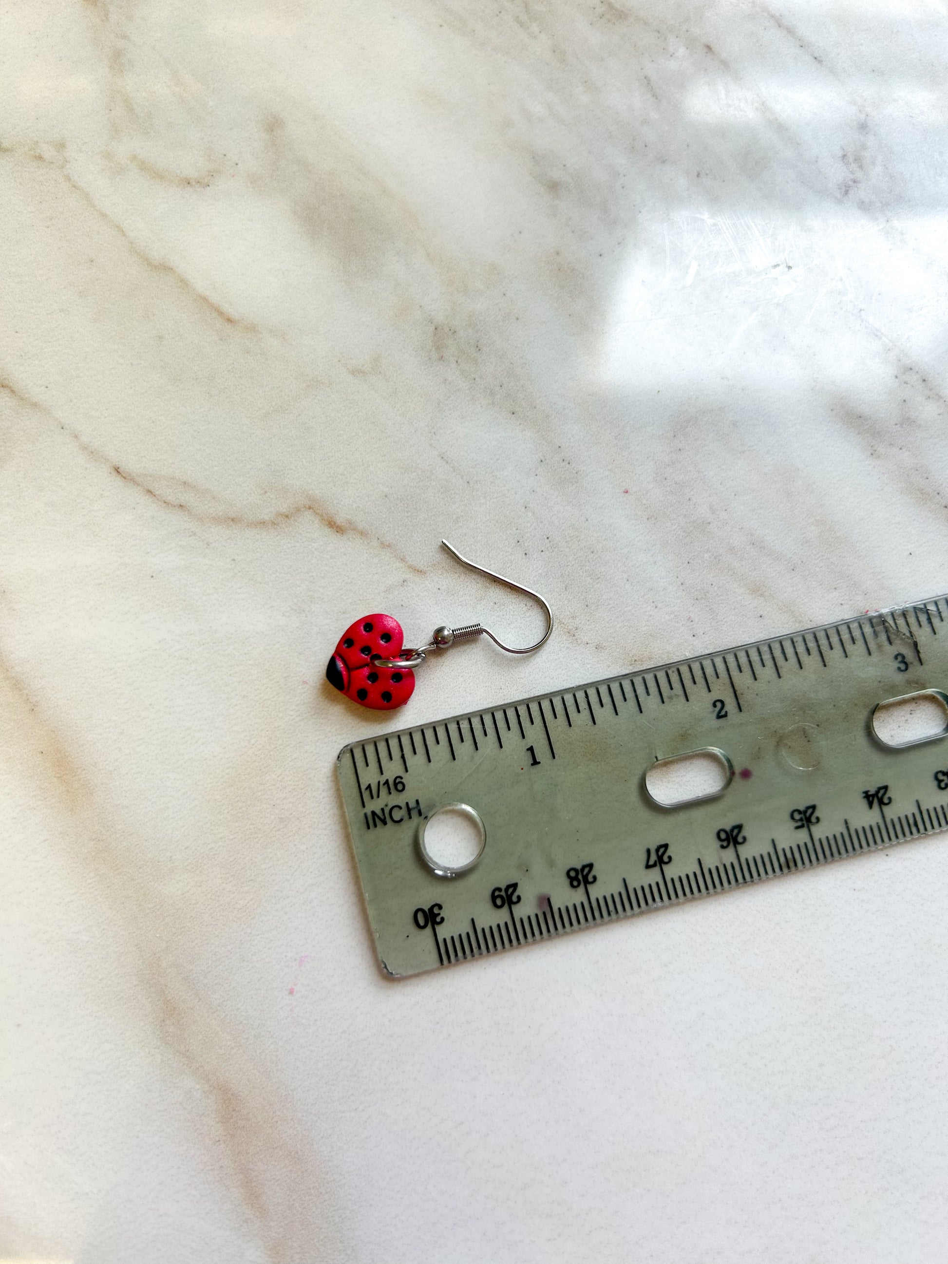 Heart shaped earrings that are painted to look like ladybugs, laying next to a ruler to demonstrate that they are approximately 1 inch in length ￼