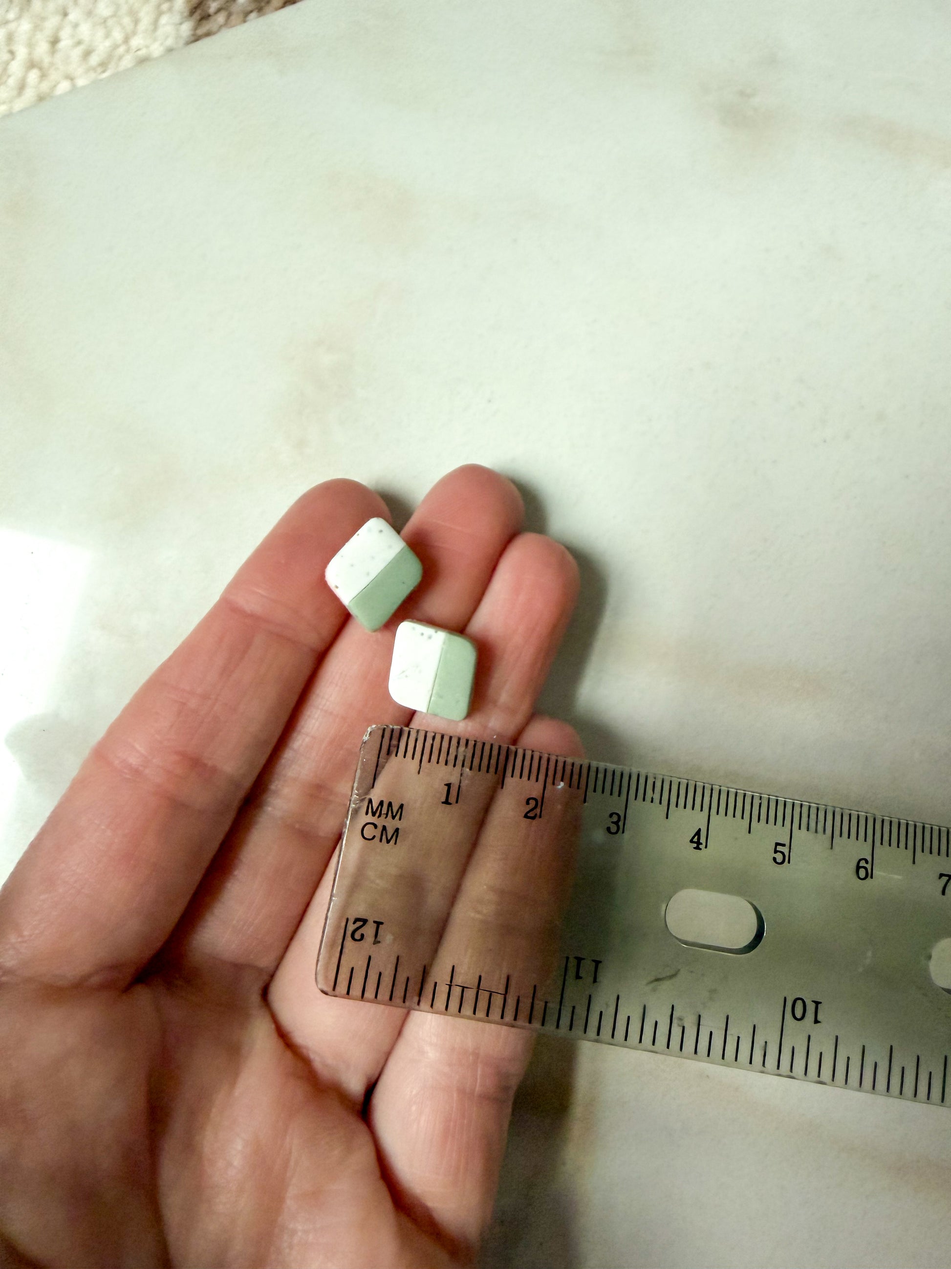 Green and white earrings held in a hand against a ruler for scale. About 1 cm long