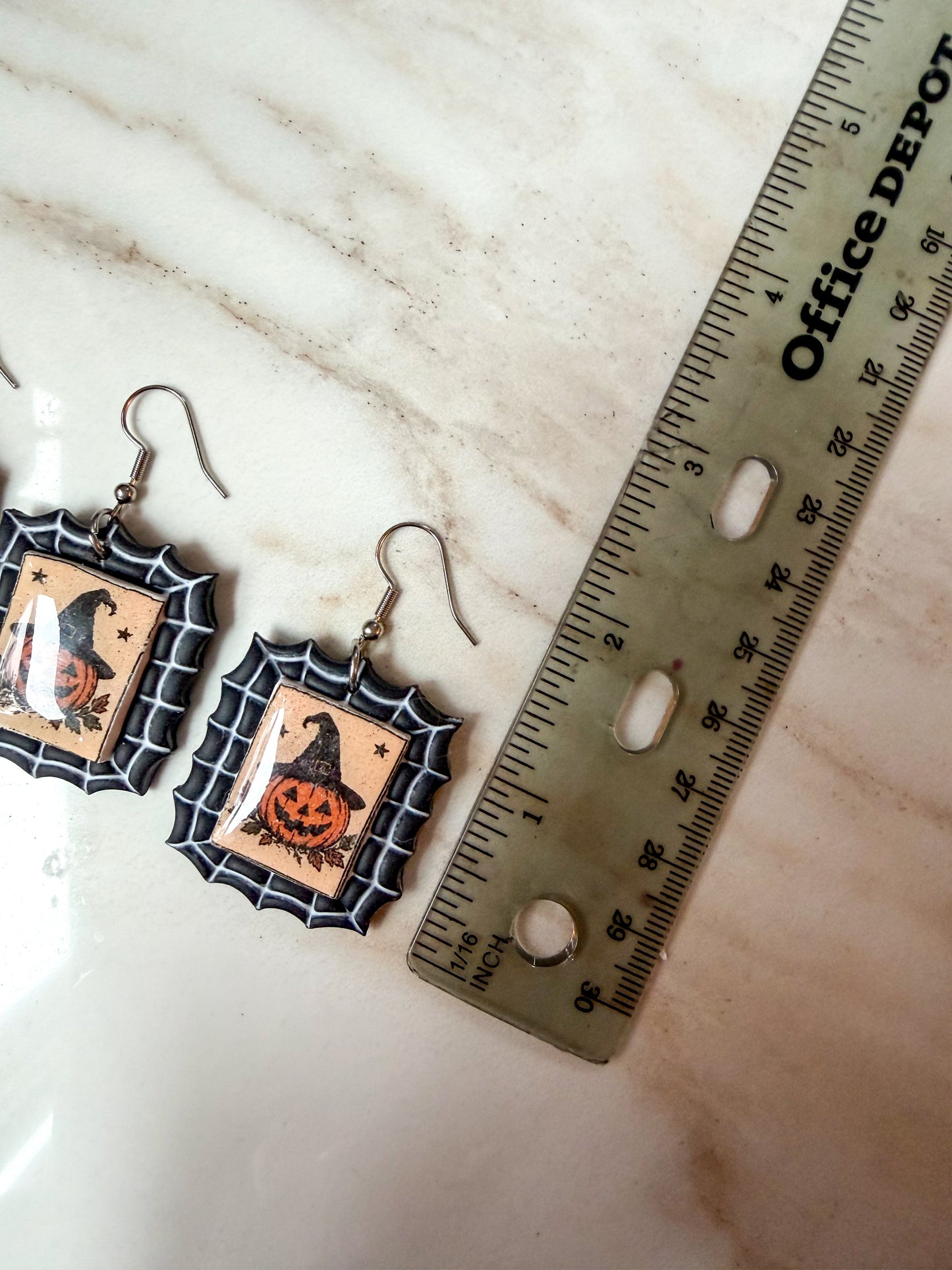 A pair of Halloween-themed drop earrings displayed on a light marble surface next to a ruler for scale. About 2 inches in length. 