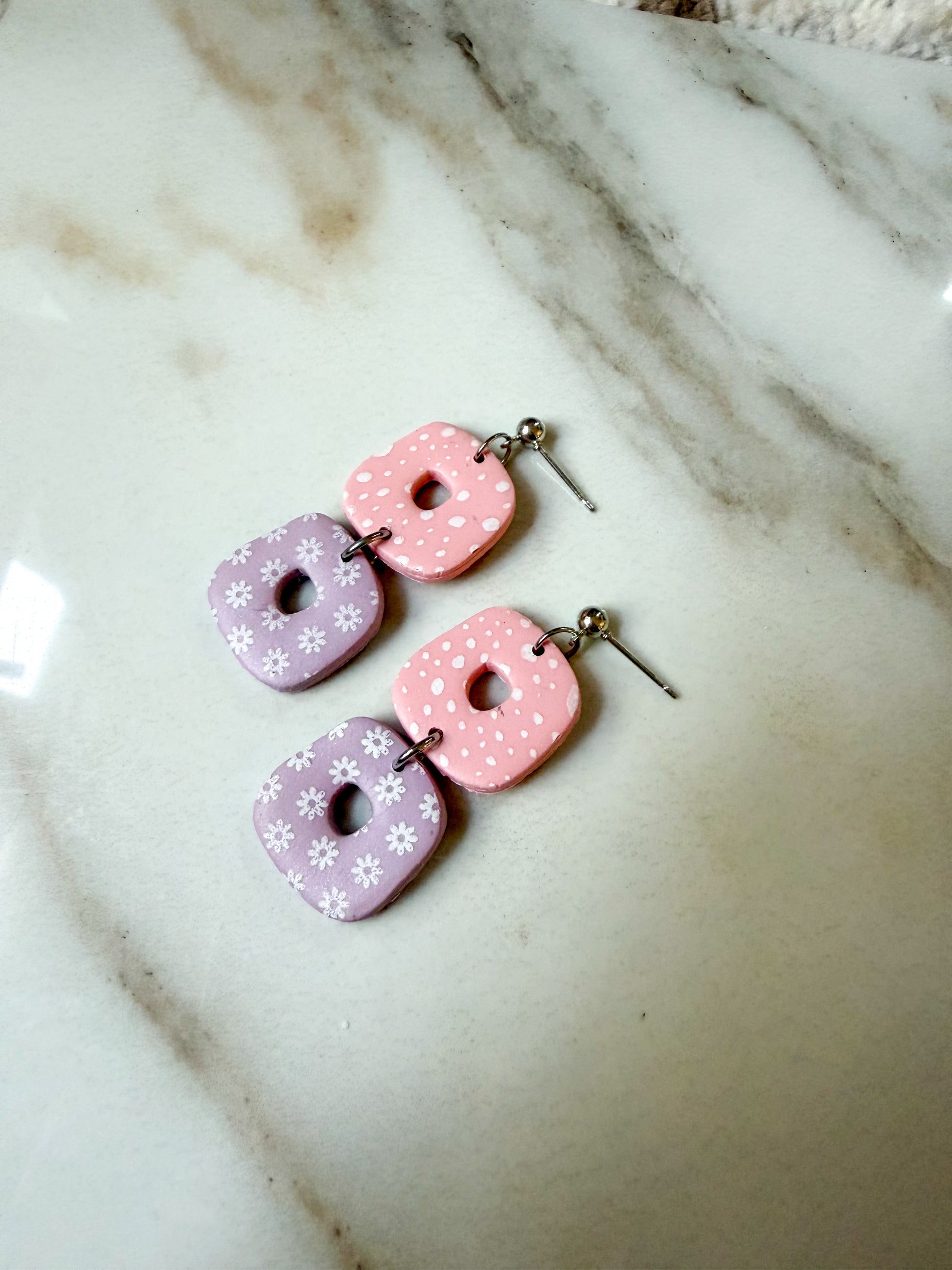 A pair of handmade dangle earrings with pastel pink and lavender donut-shaped charms decorated with white polka dots and floral patterns, resting on a white marble surface.