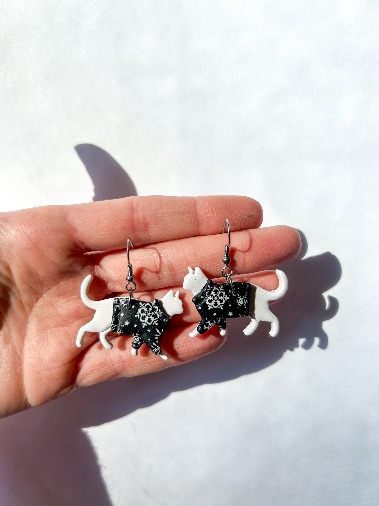 Hand holding two white cat-shaped earrings against a white background. The cats have Black snowflake sweaters. 