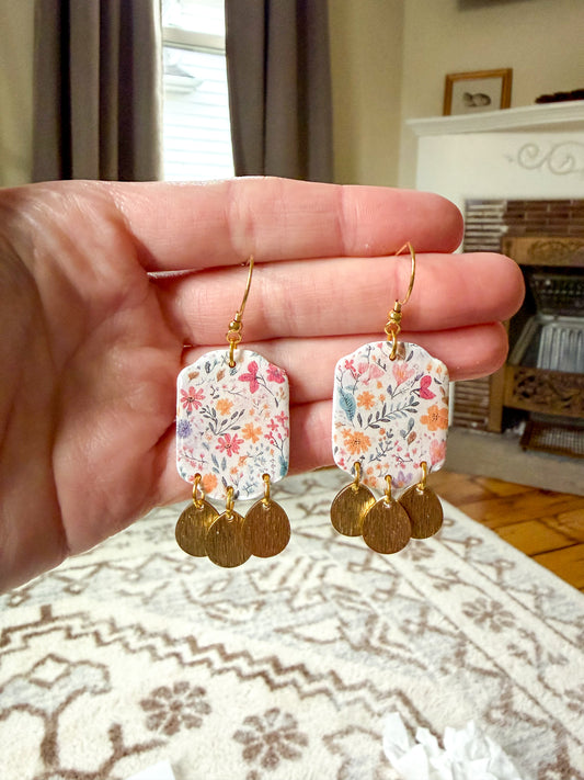 Hand holding floral earrings with gold accents against a home interior background
